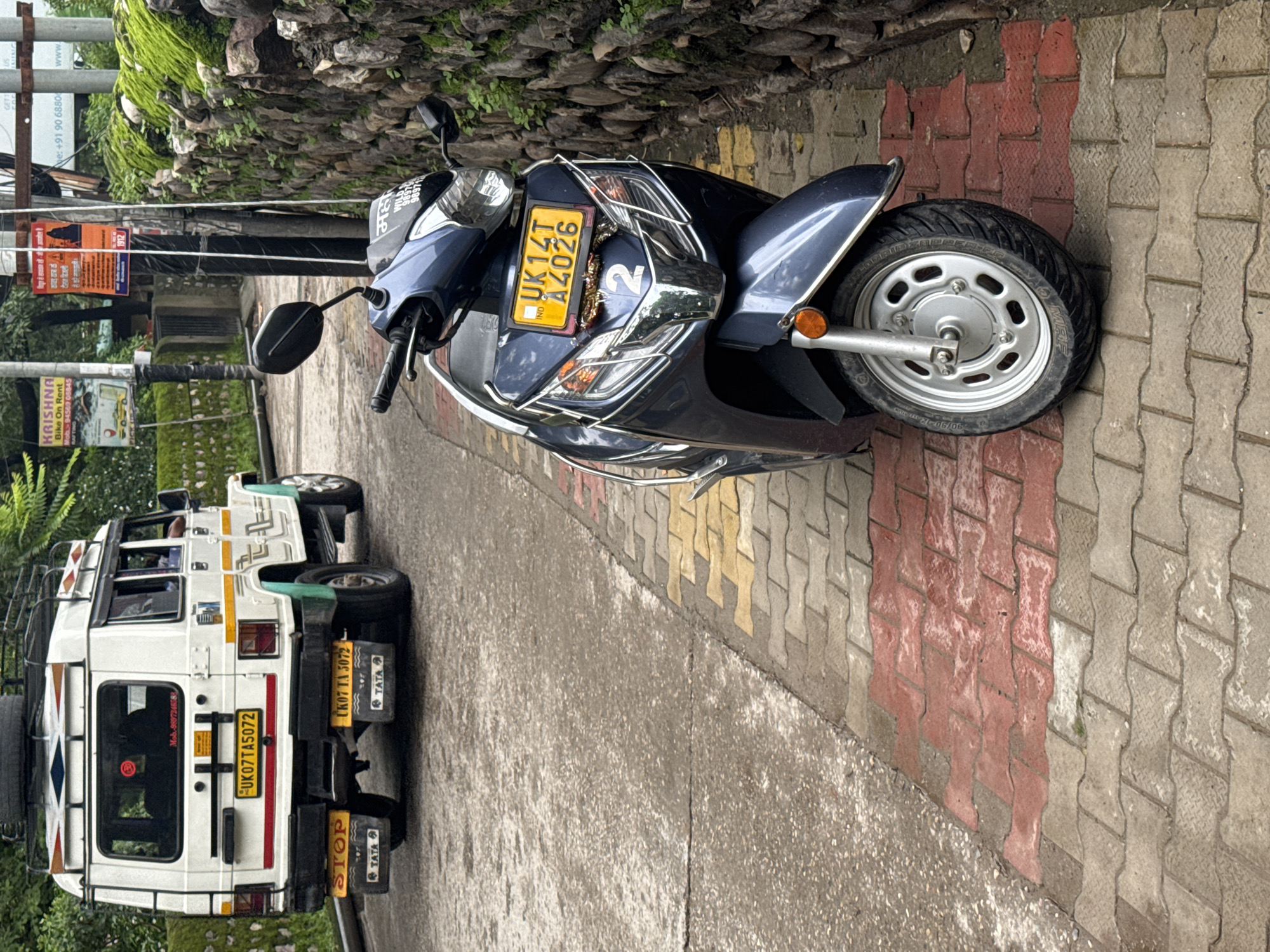 In Rishikesh, India, a dark blue scooter is parked on a brick-patterned sidewalk next to a stone wall covered in moss and vegetation. The scooter's license plate reads "UK14T A4026". To the left of the scooter, the rear of a white van is visible, parked on a paved road. The van has a roof rack, a spare tire mounted on top, and multiple license plates and stickers on its back. One license plate on the van reads "UK07TA5072". The environment appears to be an outdoor street scene with lush greenery in the background.