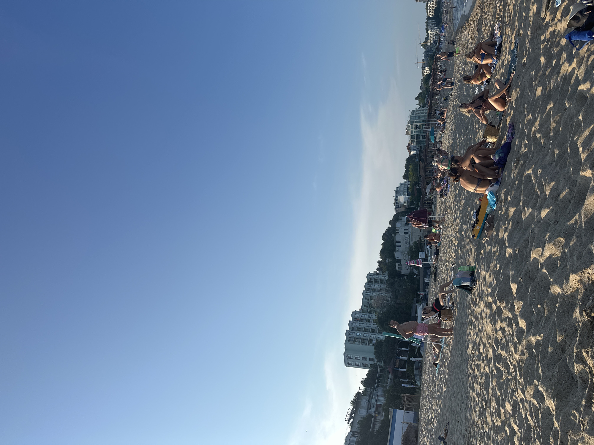 The image captures a busy beach scene with people sunbathing and relaxing on the sand. In the background, several multi-story buildings are visible on a hillside overlooking the beach, suggesting an urban coastal location. The sky is a clear, bright blue with a few wisps of clouds. The sand is a light brown, marked with shadows from the sunlight, indicating it is likely late afternoon or early morning. Several individuals are scattered across the beach, some lying on towels or beach chairs, while others are seated in groups. Beach umbrellas and bags are also present. The overall atmosphere appears to be one of leisure and recreation on a warm day.