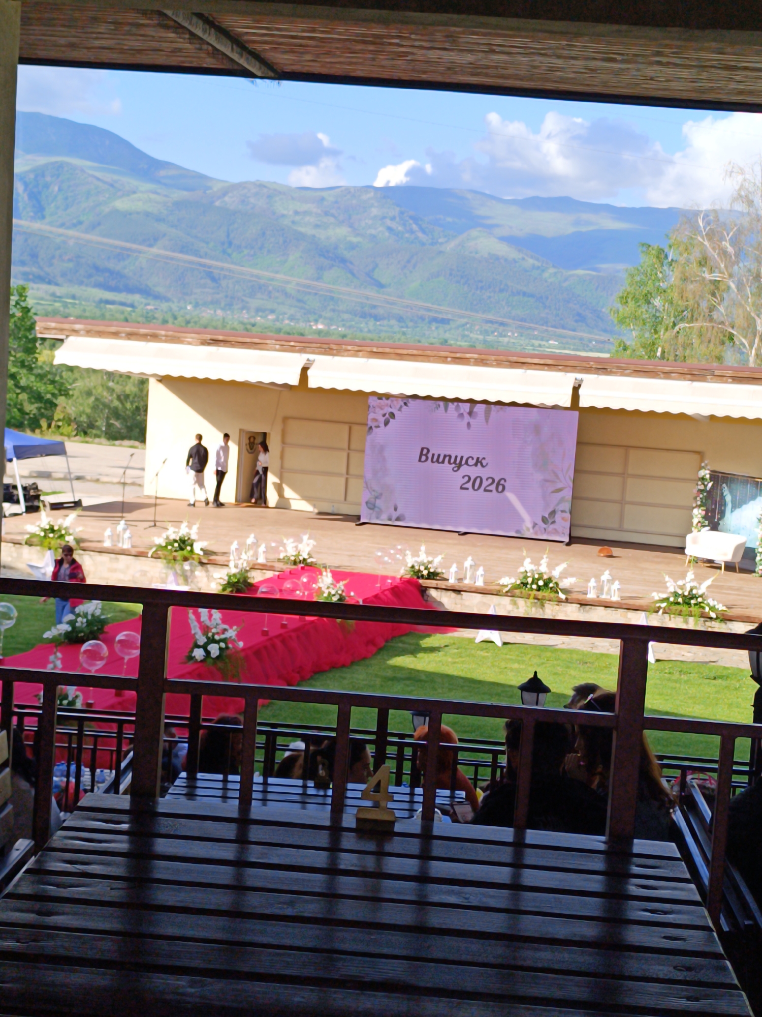 The image shows an outdoor graduation ceremony in Chelopech, Bulgaria. The event is set on a lawn in front of a tan building. A red carpet leads to a stage adorned with white floral arrangements. A large banner displays the words "Bunyck 2026". People are visible on the stage and surrounding area. A table and railing are in the foreground, suggesting the photo was taken from an elevated patio or balcony. Mountains are visible in the background.