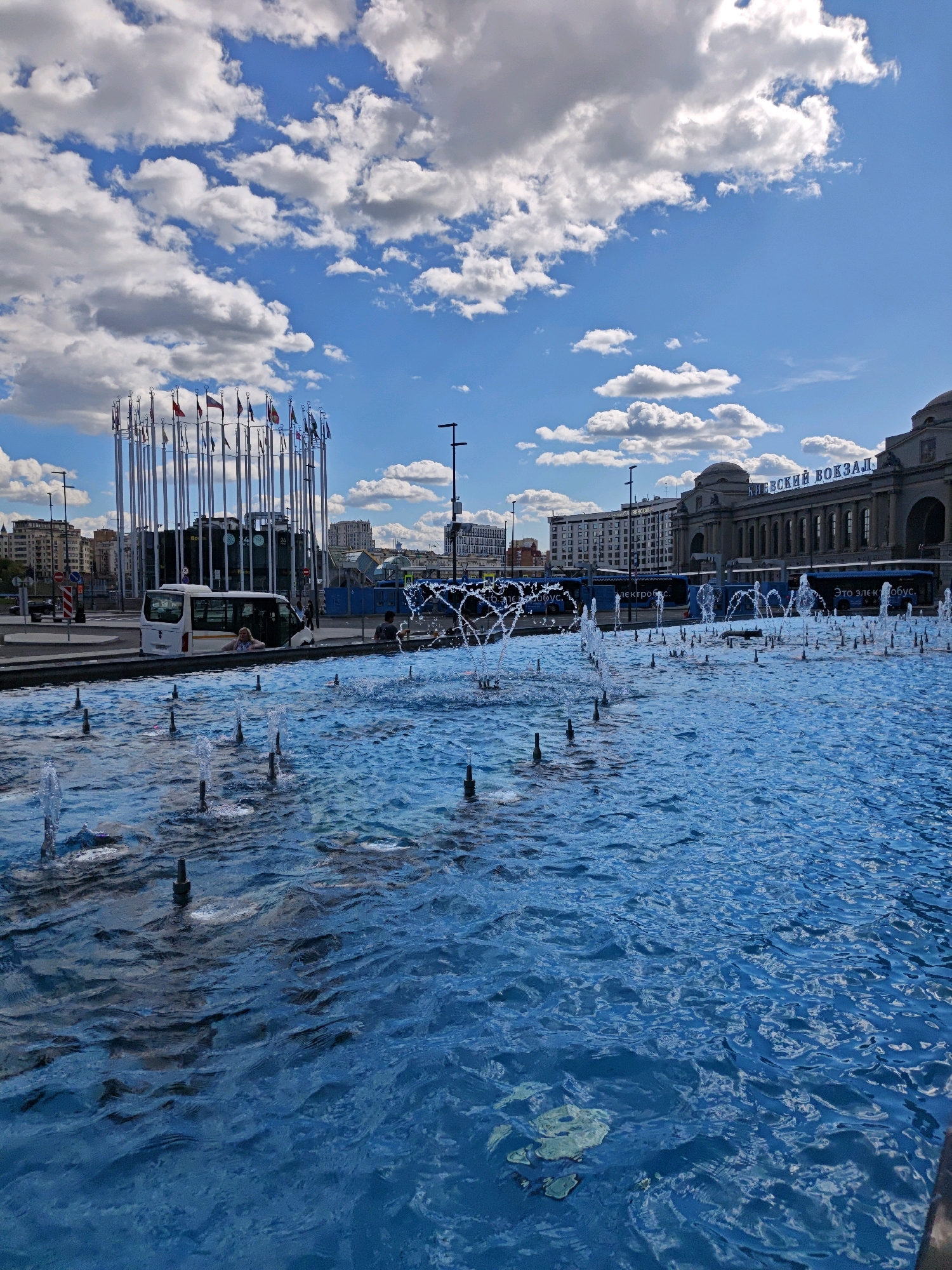 The image depicts a wide-angle view of a bright, outdoor urban scene in Moscow, Russia. In the foreground, a large, vibrantly blue fountain dominates the lower half of the frame, with numerous water jets spraying upwards. The water is rippled, suggesting a gentle breeze.
Behind the fountain, on the left, is a circular structure adorned with many flags of various nations. A white bus is parked nearby, with a person leaning out of the open side door. Further in the background, a large, classical-style building with arched windows and domes is visible, bearing Cyrillic lettering that reads "КИЕВСКИЙ ВОКЗАЛ" (Kievsky Vokzal, meaning Kiev Station). Buses, possibly public transportation, are lined up in front of the station. The sky above is a clear blue, punctuated by scattered white, fluffy clouds. The overall impression is of a public square or plaza, likely a transportation hub, on a sunny day.
