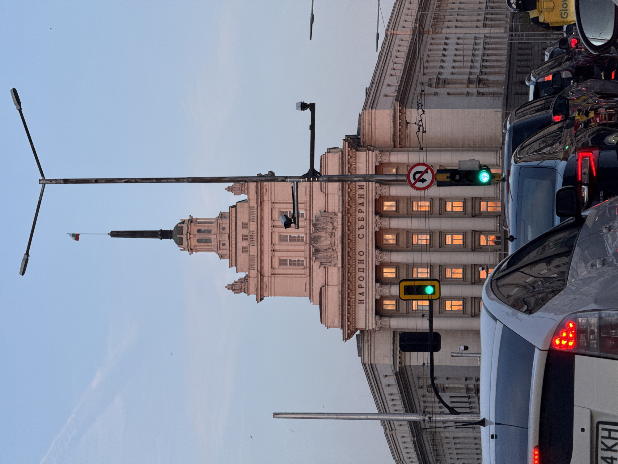 An eye-level shot captures a busy street scene in Sofia, Bulgaria, at what appears to be dusk or early evening. The prominent subject is a grand, classical building, identified as the National Assembly (Народно събрание) by the inscription on its facade. The building features multiple stories, ornate details, and a tall central tower or spire topped with a small Bulgarian flag. Many of its windows are illuminated with warm, orange light, contrasting with the pale blue sky.
In the foreground, a line of cars is visible, indicating traffic. The closest vehicle is a white car, showing part of its rear with illuminated red taillights and a license plate fragment ending in "4 KH". Reflections of the building and other vehicles are visible on its surface. Other cars, including a dark-colored van, are also part of the traffic jam.
A green traffic light is clearly visible, suggesting that traffic is currently allowed to proceed in this direction, despite the apparent congestion. Next to the traffic light, a red circular sign with a diagonal line over a U-turn arrow indicates "No U-Turn." Streetlights and overhead wires are also part of the urban landscape. In the upper right corner, a yellow "Glovo" delivery bag or box is reflected in a car mirror. The sky is largely clear, with a faint contrail and a tiny, distant object, possibly an aircraft or bird, discernible. The overall atmosphere is one of an active urban setting during the transition from day to night.
