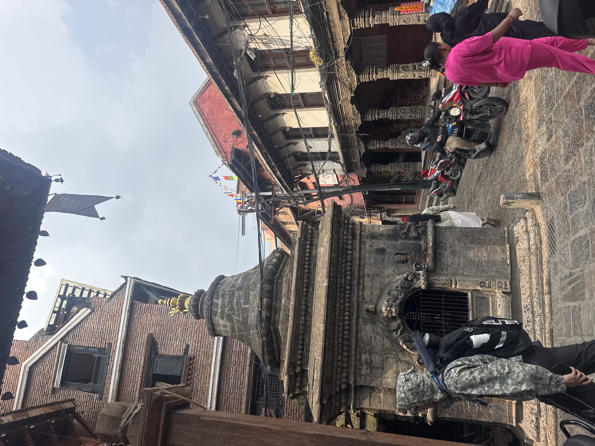 The image depicts a street scene in Lalitpur, Nepal, showcasing a historical structure, possibly a temple or monument, with intricate stone carvings and details. The structure is flanked by buildings constructed with brick and timber, displaying a blend of traditional Nepalese architecture and modern elements. Overhead, a dense network of power lines and cables create a canopy above the street.
Several individuals are present in the scene. Two are wearing pink and black attire and are near parked motorcycles on the right side of the image. One individual is lying on the ground in white attire. Another person is lying on the ground wearing camouflage gear.
The architectural details, presence of people, and utility lines suggest a bustling urban environment rooted in historical tradition. A black flag flutters on the left of the image, and prayer flags stretch across the scene, contributing to the location's cultural context.