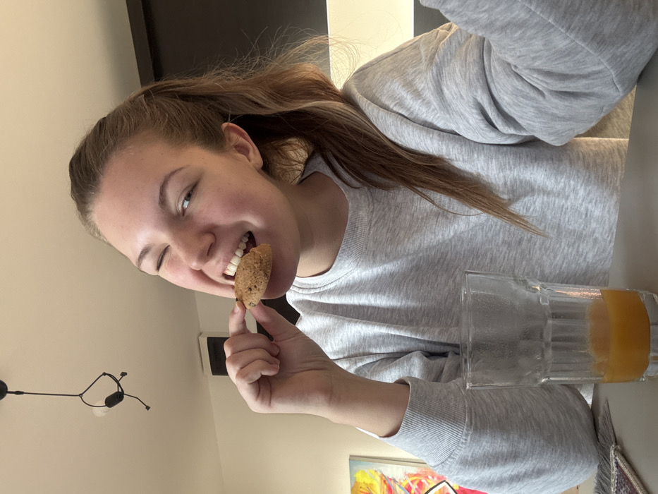 A young adult female is depicted indoors, in the city of Vozdovac Urban Municipality, Serbia. She is seen from the chest up, positioned centrally, with her head slightly angled and her left eye closed in a wink, looking towards the viewer. Her mouth is open, biting into a circular chocolate chip cookie, which she holds with the thumb and index finger of her right hand. She is wearing a light grey long-sleeved crew-neck sweatshirt. To her lower left, a clear, ribbed drinking glass containing an orange-colored liquid, likely juice, rests on a smooth grey surface. A small, patterned textile placemat or coaster is partially visible beneath the glass. The background consists of a light-colored ceiling with a black, multi-armed pendant light fixture hanging from it in the upper left. A white rectangular object, possibly an air vent or speaker, is mounted on a light-colored wall in the mid-left. A colorful abstract artwork is partially visible on the far left, and a dark roller blind is visible on the far right. The scene is brightly illuminated.