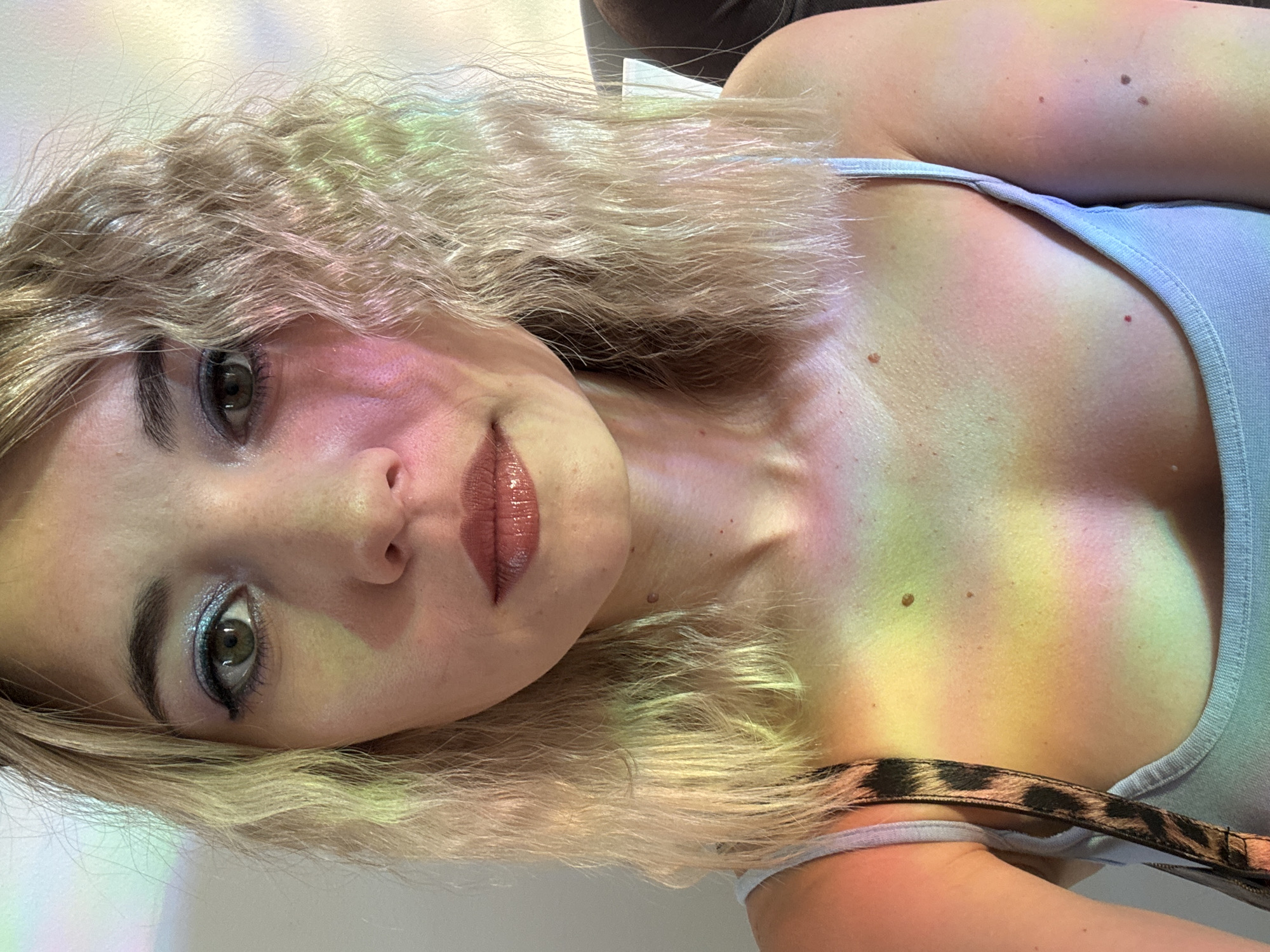 A close-up, frontal, waist-up selfie of a fair-skinned blonde woman with wavy hair, taken indoors in what appears to be an artificially lit environment. She is wearing a light blue tank top. Her face is illuminated by a colorful light display casting hues of pink, yellow, and green across her skin, hair, and clothing. She is wearing eye makeup with blue and glitter tones, and lip gloss. Her head is slightly tilted to her right. The background is indistinct and appears to be a wall with some light wash.