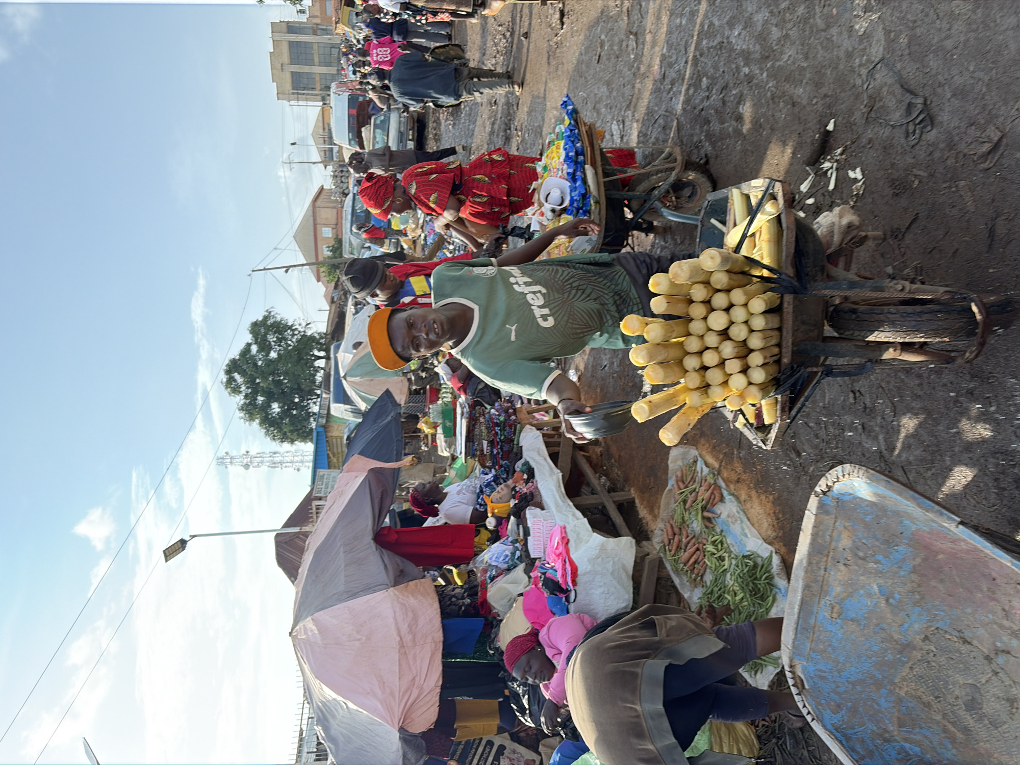 The image captures a bustling market scene in Gura Top, Nigeria, bathed in natural sunlight. In the foreground, a man wearing a green t-shirt and an orange baseball cap stands behind a wheelbarrow filled with cut sugarcane. His gaze is directed towards the camera. The wheelbarrow is laden with stalks of sugarcane, some cut into short pieces. To the left of the man, another wheelbarrow, predominantly blue and weathered, sits empty.
Further into the market, several vendors are actively engaged. One woman, adorned in a red and patterned dress and headscarf, is interacting with another person. In the background, various stalls are set up under awnings, displaying a variety of goods including textiles and produce. People are seen moving through the market, some carrying items. Vehicles, including what appear to be minibuses and cars, are present in the background, suggesting the market is located on or near a road. The ground is a mix of dirt and debris, indicative of an outdoor market environment. The sky above is clear with some wispy clouds.