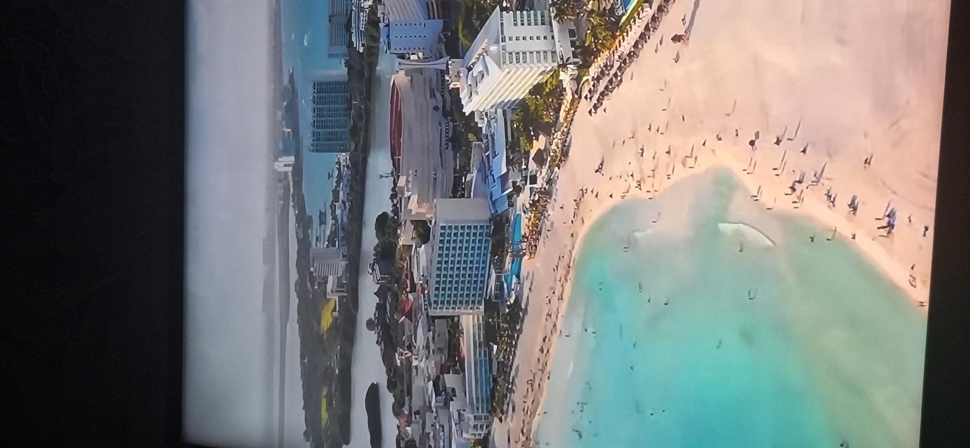 An aerial shot shows a coastal city with several large hotels and resorts lining a sandy beach. The ocean is a vibrant turquoise color, with people swimming and sunbathing on the shore. Several boats are visible in the water. In the background, there are more buildings and lush greenery, suggesting a tropical or subtropical location. The scene is bathed in bright sunlight, creating long shadows on the beach. There is no visual information in the image to suggest it is located in Bila Tserkva, Ukraine.