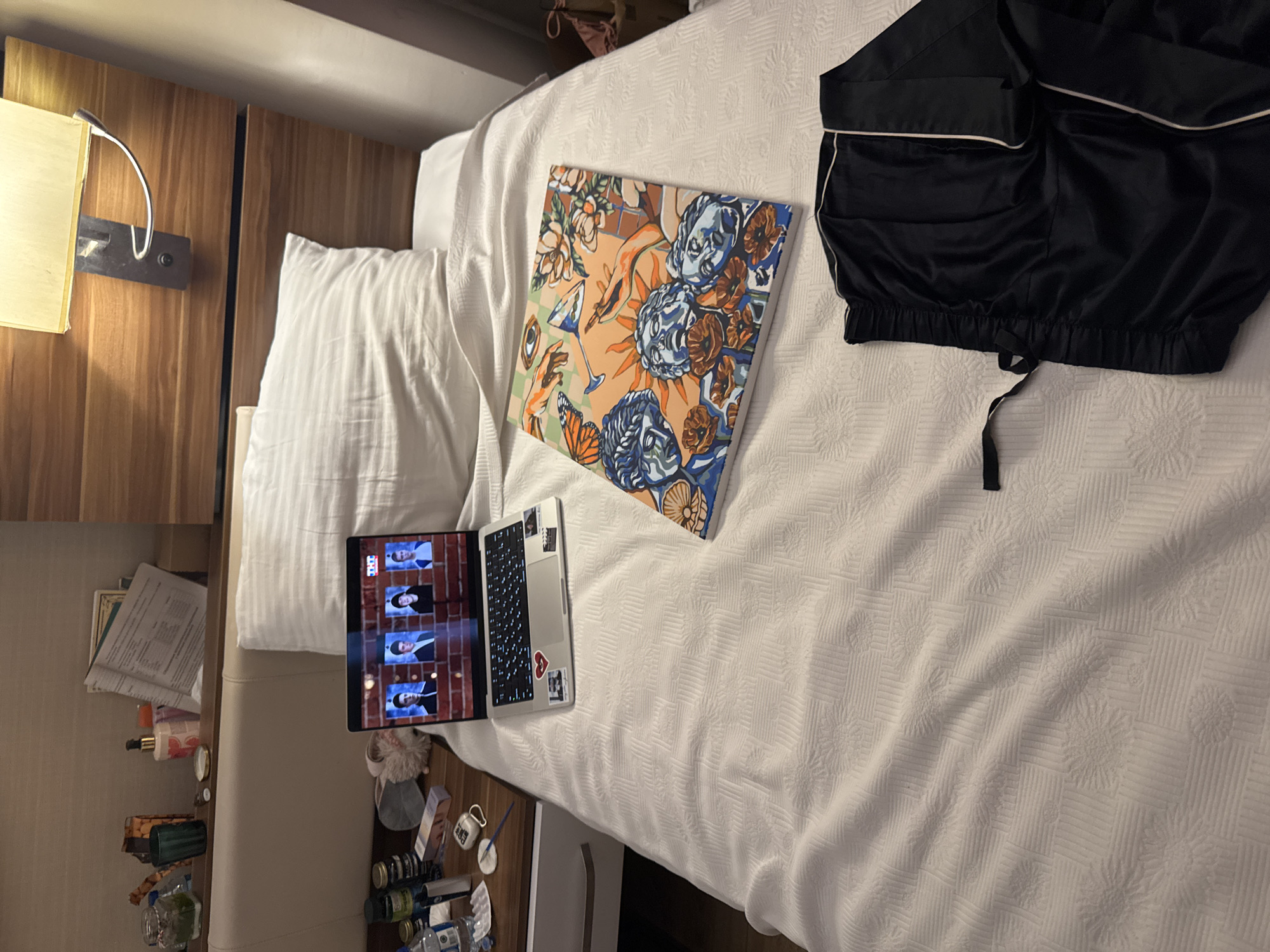 The image depicts a hotel room with a bed, a nightstand, and a wall-mounted lamp. On the bed, there is a laptop displaying a video call with four participants, a painting, and a pair of black silk pajamas. The nightstand holds various items, including bottles of water, medication, and toiletries. The overall scene suggests a person is relaxing in their hotel room, possibly preparing to sleep or unwind after a long day. The city of Aksu, Turkey is not discernible from the image.