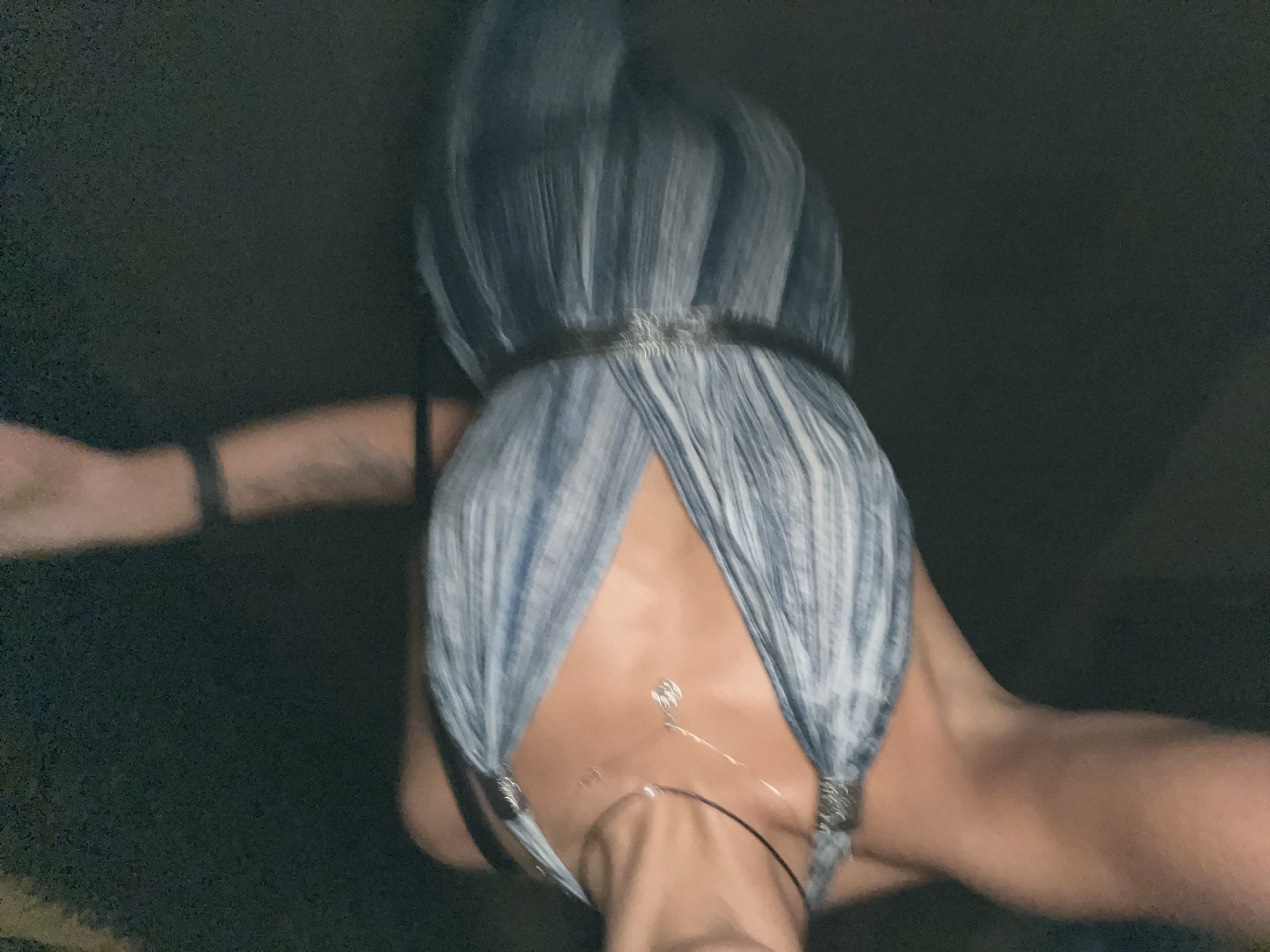 The image shows a person with light skin, wearing a grey and white striped garment that covers their chest and shoulders. The garment is gathered at the top, possibly forming a type of headdress or head covering. A dark band is visible around the top of the head covering. A necklace is visible on the person's chest. The background is dark and blurry, with some indistinct light areas. The person is slightly out of focus. There is a wrist watch on the left arm. The location is not discernible from the image. The city of Unknown, Indonesia is specified, but there is no way to verify this location.