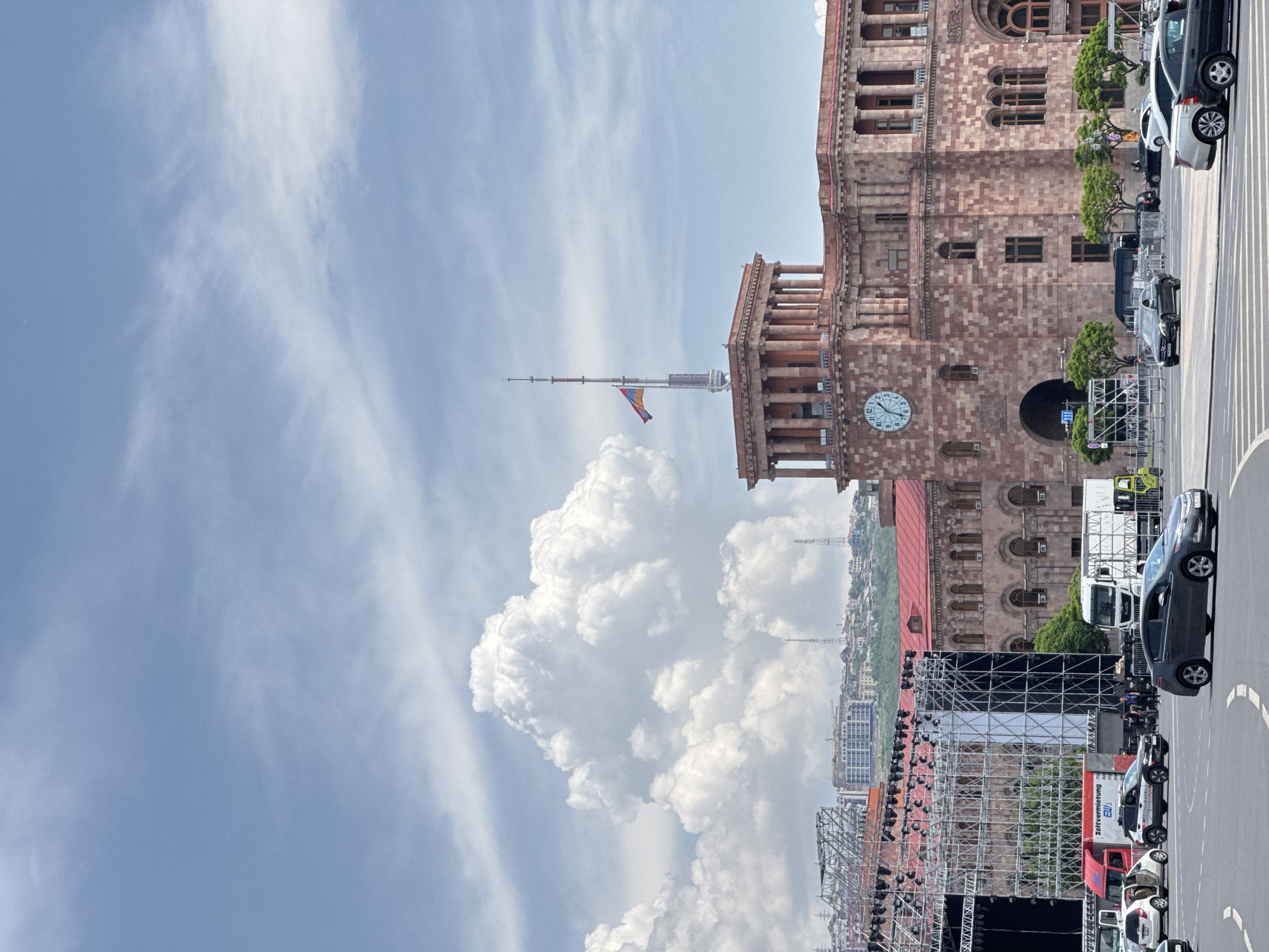The image depicts a scene in Yerevan, Armenia, focusing on the Republic Square area. The most prominent feature is the Government House, a large stone building with a clock tower topped by a flagpole flying the Armenian flag. The photograph captures a stage setup, possibly for an event or concert, is in the foreground. Cars and some pedestrians are visible on the street. The sky is filled with clouds, and other buildings are present in the cityscape.