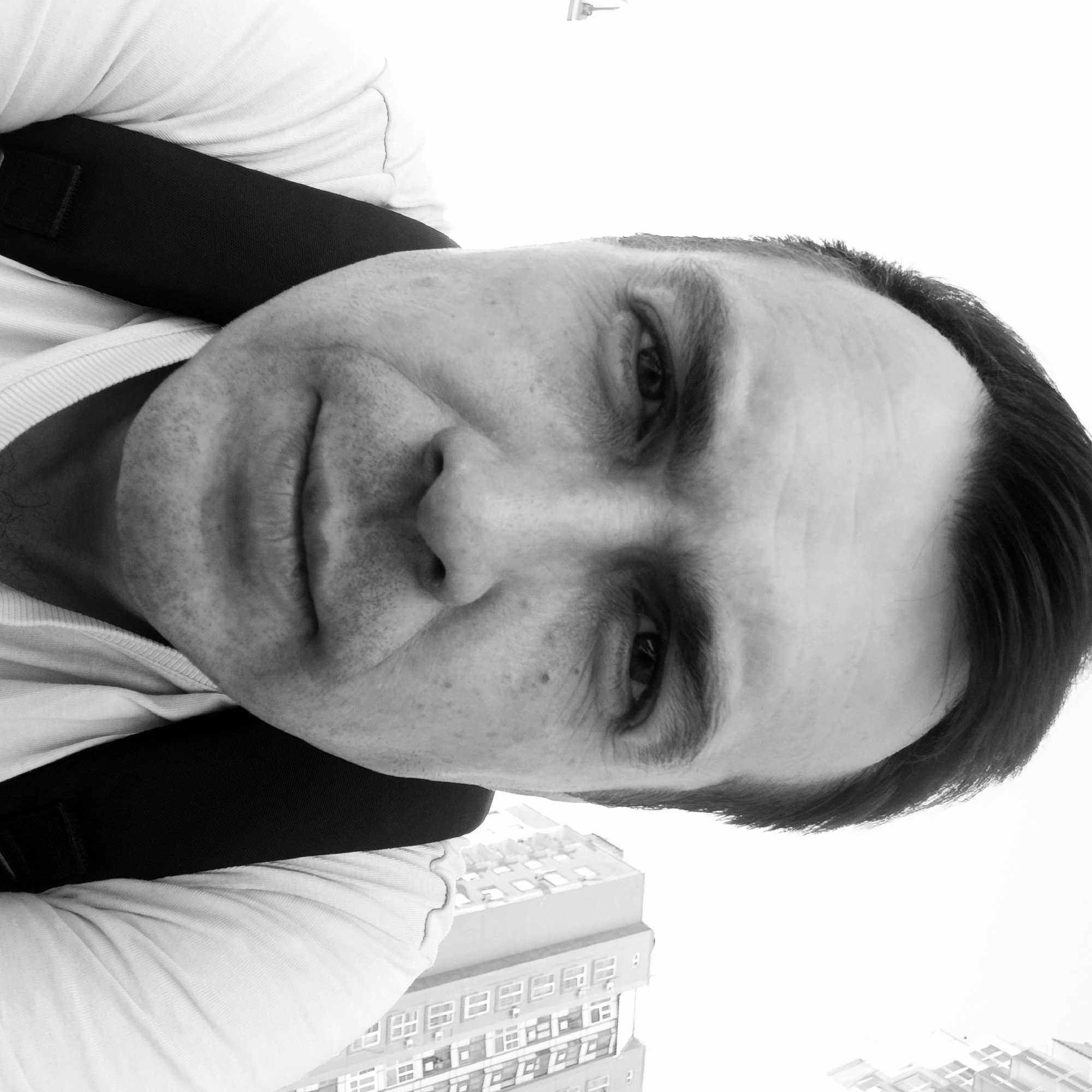 A black and white, close-up, low-angle selfie shows a man wearing a white t-shirt and a backpack. He has dark hair, fair skin with some visible pores and stubble, and is looking directly at the camera with a neutral expression. The background is out of focus, showing a bright sky and the side of a building with multiple windows. The image was taken outdoors, likely in an urban environment.