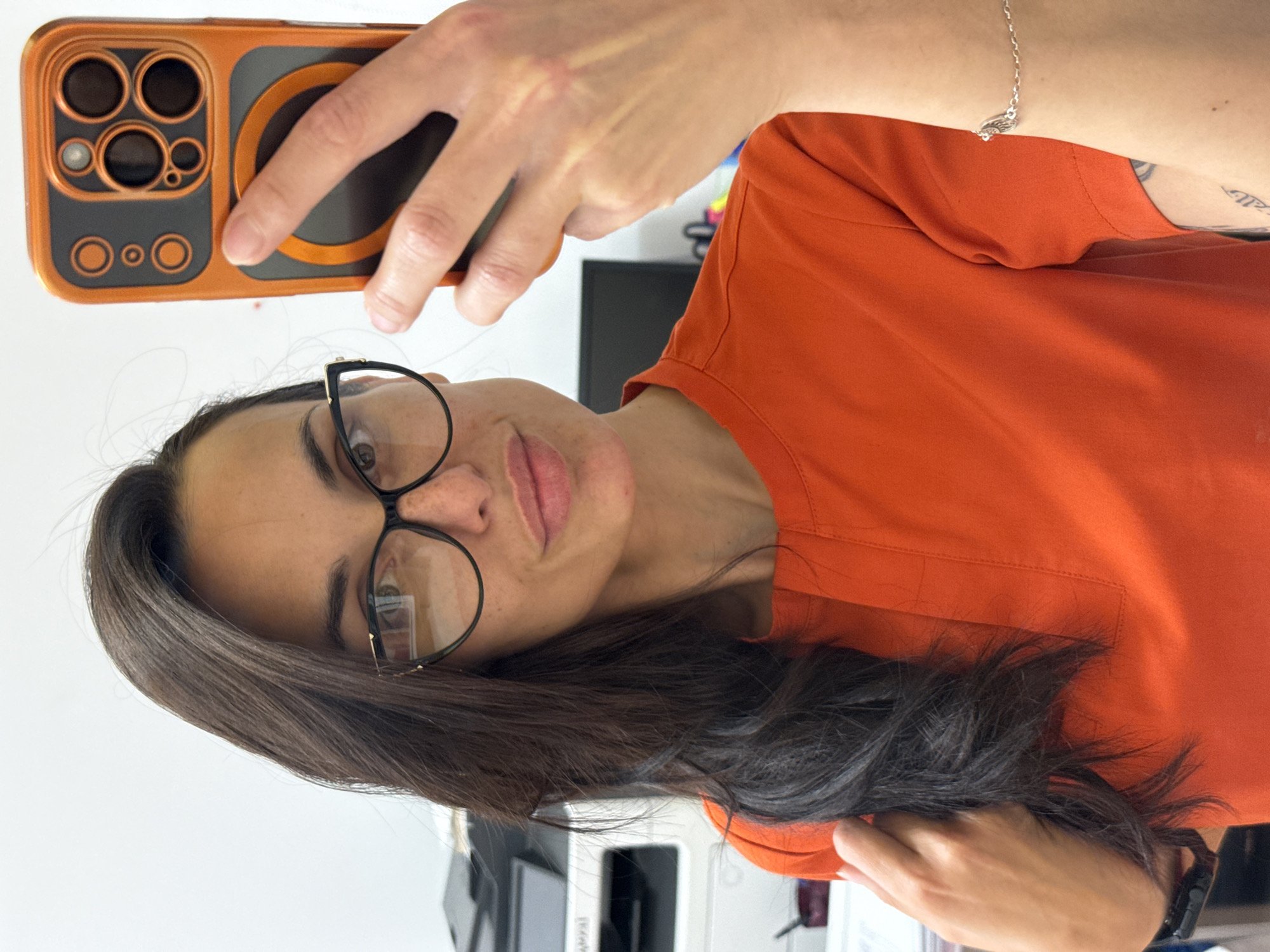 A woman is taking a selfie with an orange smartphone. She is wearing an orange shirt and glasses. A silver bracelet is visible on her left wrist. The location cannot be determined.