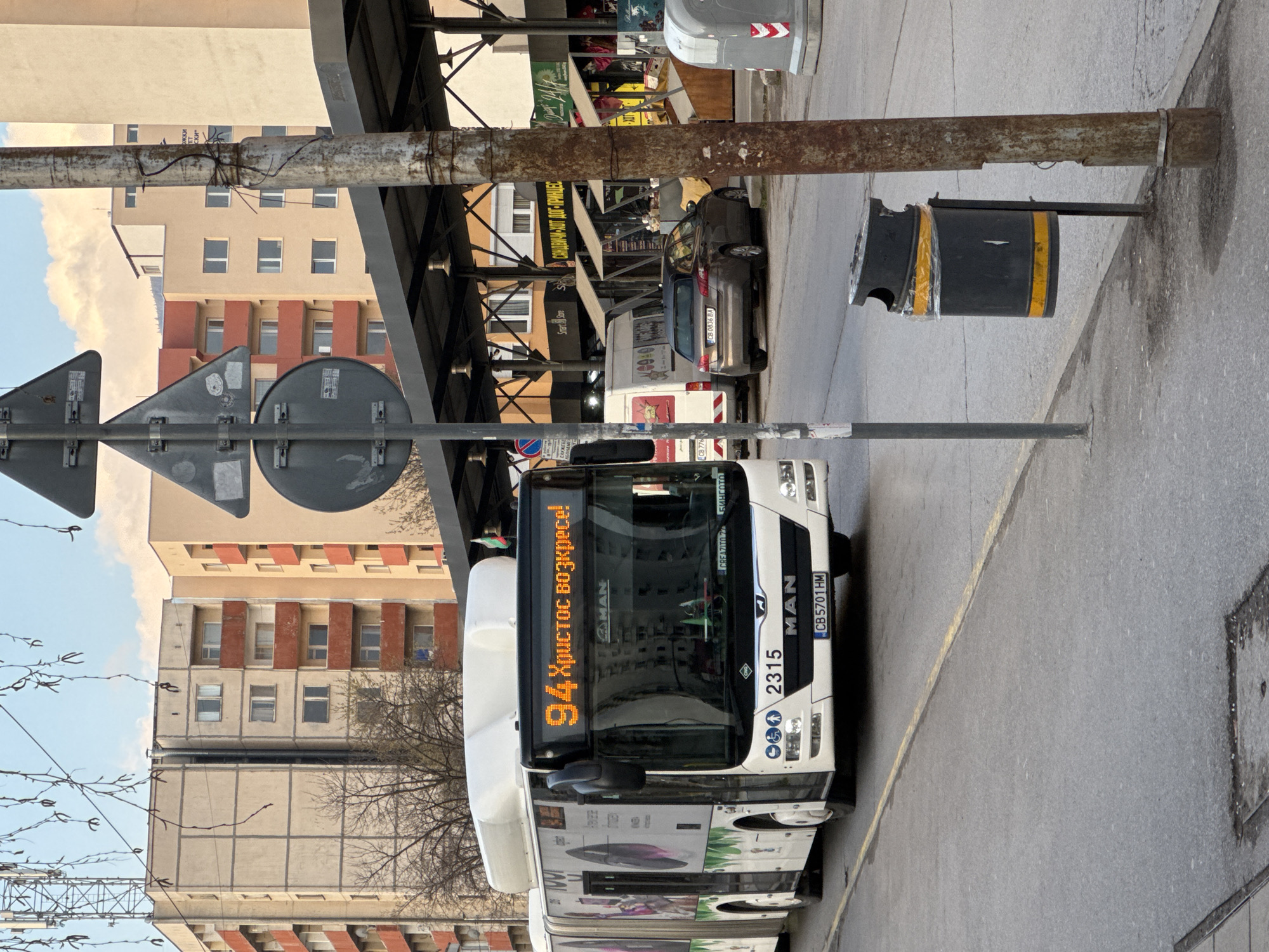 The image depicts a street scene in Sofia, Bulgaria. A white MAN bus with license plate CB 5701 HM, operating route 94 ("Христос возкресе!" displayed, meaning "Christ is Risen!"), is prominent. The bus stop includes signage, a metal structure providing cover, and a damaged trash can. Parked vehicles, including cars with license plates CB 0836 BA and CB 776, are visible along the street. In the background are multi-story apartment buildings and commercial establishments with awnings, including one offering sandwiches and hot dogs. The scene appears to be in a somewhat urban and likely commercial area of Sofia.