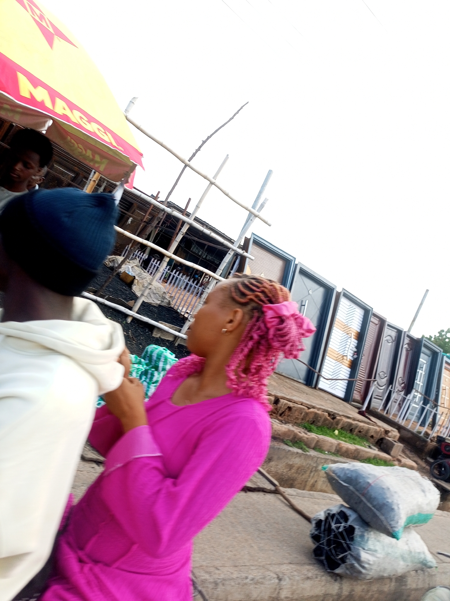 In Gombe, Nigeria, a market scene unfolds with several individuals and objects on a dusty surface. In the foreground, a person wearing a black beanie and a white hoodie is partially visible, facing away from the camera. To their right, a woman with bright pink braided hair, adorned with a pink bow, is seen in profile. She is wearing a vibrant pink dress and appears to be interacting with the person in the hoodie, her head turned towards them. Behind them, a row of metal doors, in various colors and designs, are displayed upright, suggesting a business or stall selling doors. Some of these doors have decorative patterns. A yellow awning with red accents, possibly advertising "MAGGI," is visible in the upper left corner of the image. In the background, a rudimentary structure made of metal poles and wood can be seen, along with a glimpse of other people and market activity. Piled on the ground in the lower right are two large sacks, one lighter colored with a green band and another darker, both containing what appears to be charcoal or similar material. A few dark cylindrical objects, possibly pipes or bundled materials, are also on the ground next to the sacks. The overall environment appears to be an outdoor market, typical of Gombe, Nigeria, characterized by its vibrant atmosphere and variety of goods.