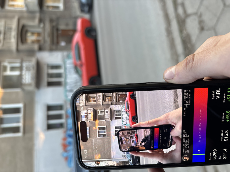 A person's right hand holds a black smartphone in portrait orientation, displaying an interactive digital interface overlaid onto a live camera feed. The phone screen shows an urban street scene in Sofia, Bulgaria, mirrored from the physical background. On the screen, a red two-door muscle car is parked on the right side of an asphalt street, with a multi-story building featuring multiple windows and small balconies behind it. Graffiti, primarily in blue and orange, is visible on a wall in the background to the left.
Within the primary phone screen, a recursive image shows a smaller smartphone held by two hands, also displaying the same scene and a person taking a photo of the red car. The phone's interface includes the time "18:03", a user profile ("valentin.georgoushki") with location "Sofia, Bulgaria", and descriptive tags "Curiosity, Intrigue Urban, Modern Observation". Below the visual feed, a horizontal gradient bar with a "+0" indicator and the prompt "Swipe / click to stake" suggests an interactive application. Financial data is displayed at the bottom: "Volume 5,269", "Symbol VIRL", "TUL $21.4", "Principal $15.8", "Revenue +$5.6", and "Creator +$1.13".
The physical background, out of focus, precisely matches the scene depicted on the phone screen: the same red car, the same building facade, and the same street environment. The ground consists of concrete pavement and an asphalt road.
