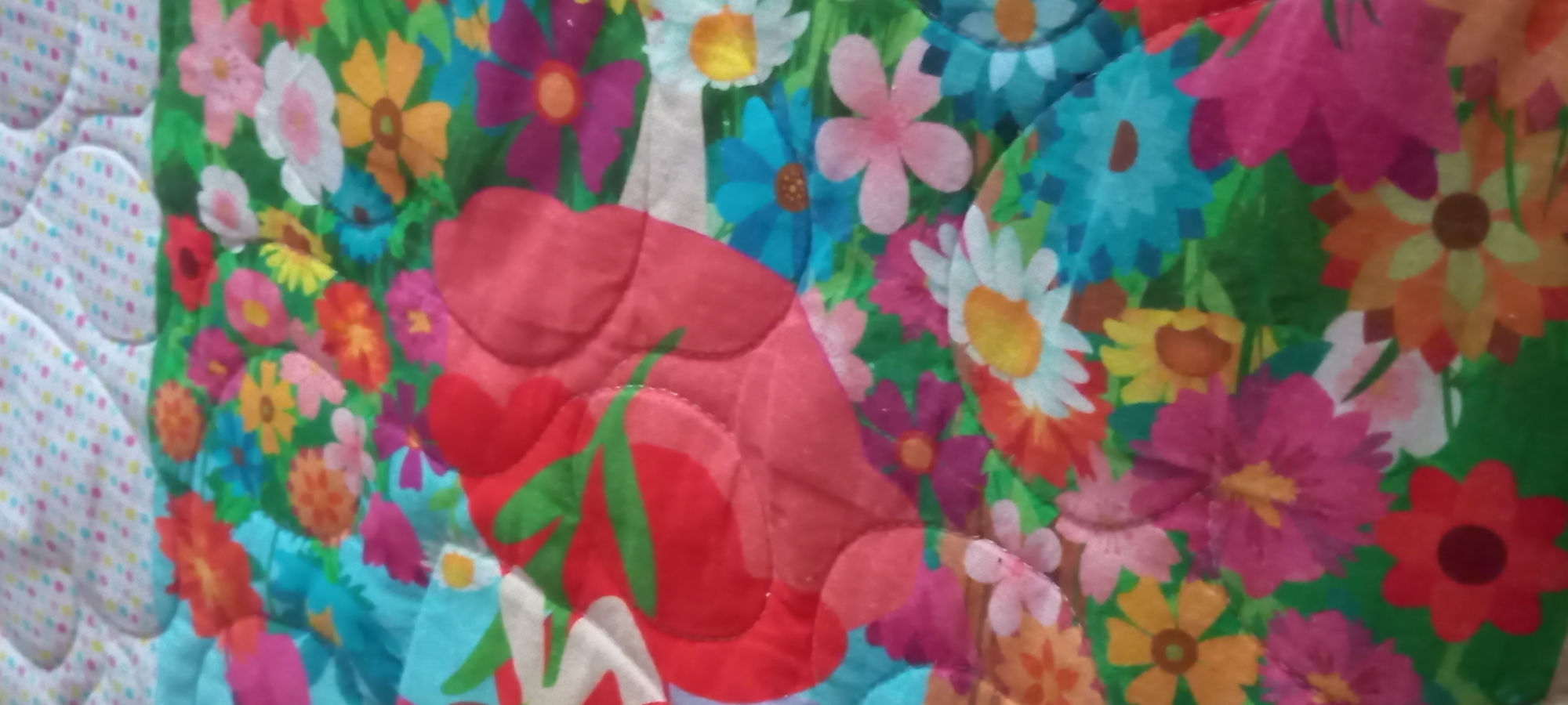 The image displays a close-up, angled view of a brightly colored, quilted fabric, potentially a blanket or comforter, located in Kryvyi Rih, Ukraine. The fabric is divided into two distinct patterns. The upper portion features a white background adorned with numerous small, multicolored polka dots in hues of pink, blue, yellow, and green. This section exhibits a puffy, rounded quilted texture. The lower and central portions are dominated by a dense, vibrant floral pattern set against a green background. This floral design includes stylized flowers of various sizes and shapes, depicted in a broad spectrum of colors such as red, orange, yellow, blue, pink, white, and purple. Prominently quilted into the floral pattern are large, red, heart-shaped forms, indicated by raised contours and visible stitching; one such heart in the center features green, stem-like details. The overall scene is characterized by high saturation and soft illumination, with no individuals, specific actions, or further environmental details discernible beyond the fabric itself.