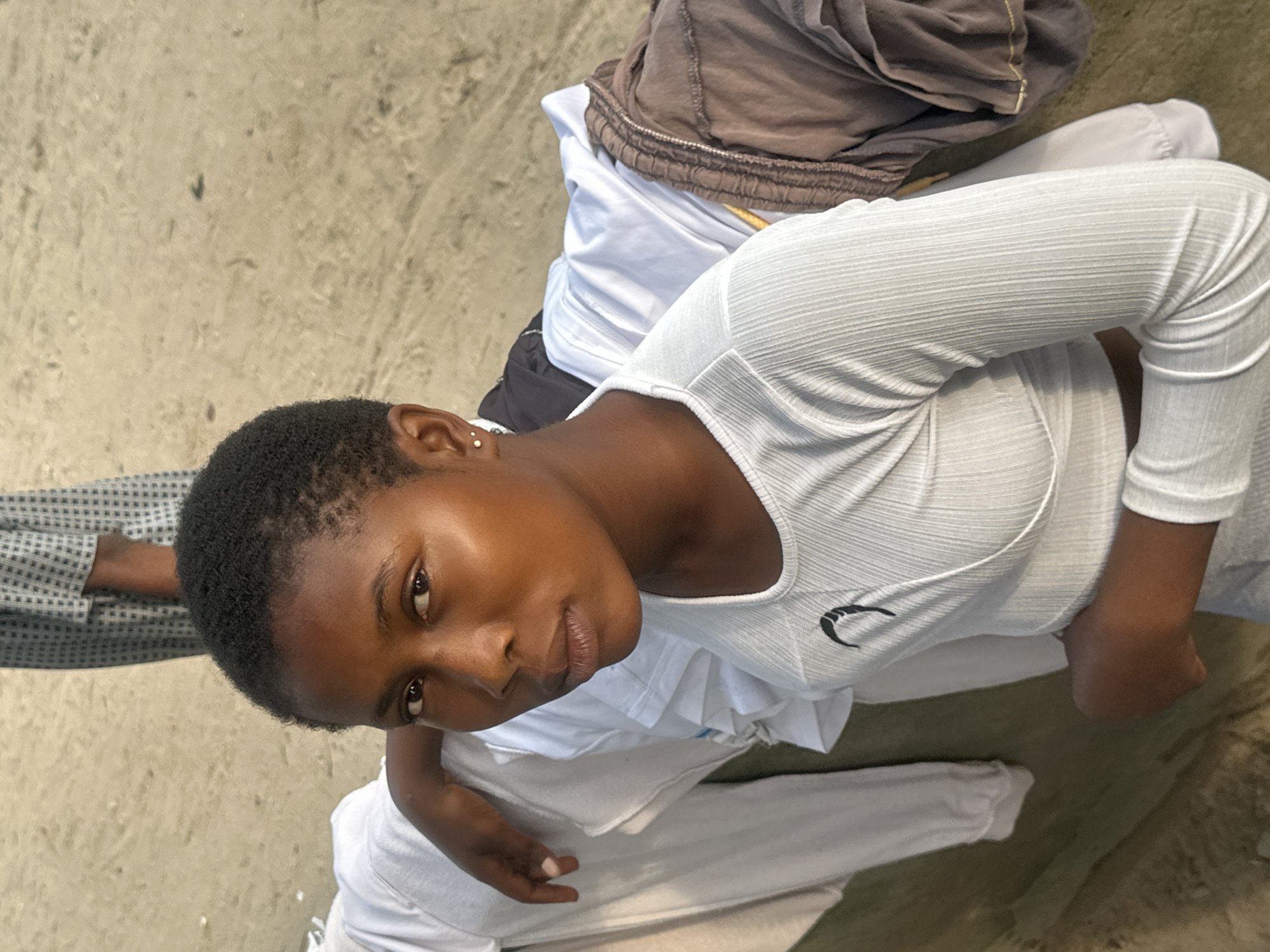 A young Black woman with short, dark hair and light brown skin is in the foreground of the image, looking directly at the camera. She is wearing a white, long-sleeved, ribbed crop top with a black crescent moon logo on the left side of her chest. Her right arm is bent and rests on a pile of clothing. She has small, gold stud earrings in both ears.
Behind her, the scene appears to be outdoors, with a sandy or dusty ground surface visible. To the left of the woman, there is a pile of white clothing, possibly laundry. To her right, there are other items of clothing, including a brown pair of pants with a drawstring. In the background, another person is partially visible, wearing a grey and white patterned garment. The overall impression is of a casual setting, possibly a beach or a public space where laundry is being laid out. The image was captured in the city of Badagry, Nigeria.