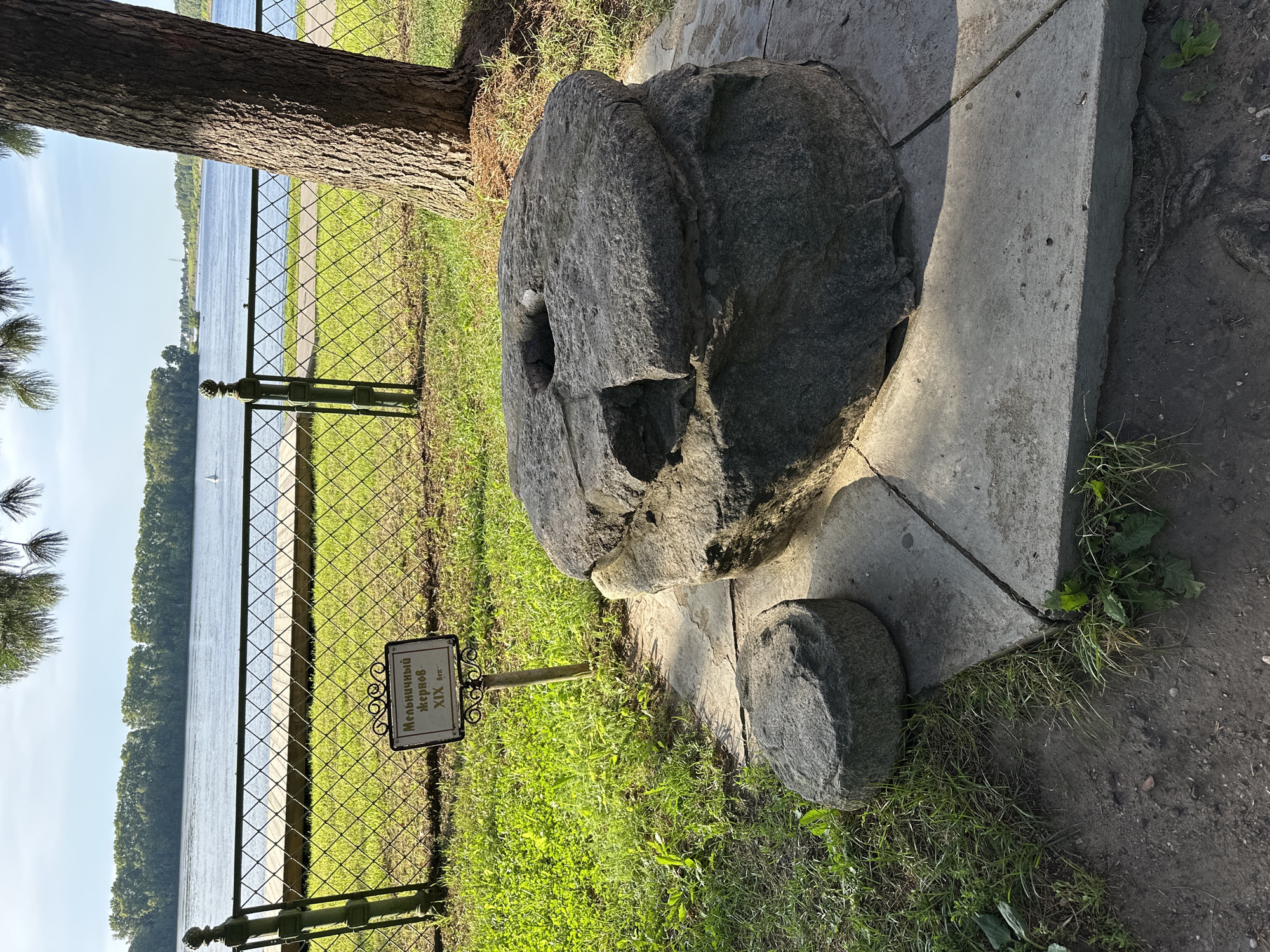 The image displays a large, circular millstone, possibly a historical artifact, placed on a stone base in an outdoor setting. To the left of the large millstone, a smaller, round stone object rests on the grass. A sign with Russian text, likely indicating the millstone's origin or historical significance, is positioned in front of the larger stone. The scene is situated near a body of water, likely a river, with a tree trunk visible to the right. The background shows a tree line and a blue sky. The location is identified as Terpilovo, Russia.