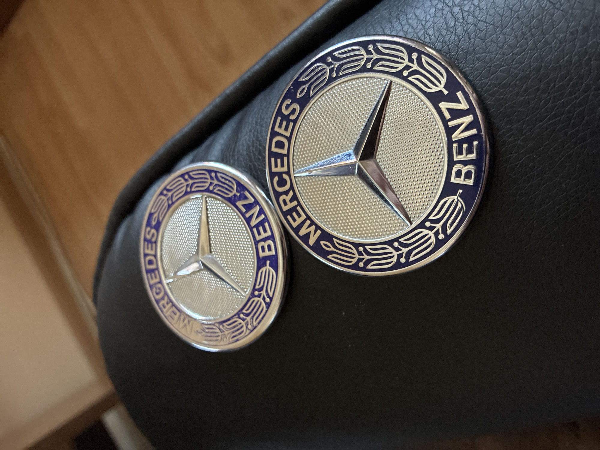 Two Mercedes-Benz emblems are prominently displayed on a dark, textured surface, possibly a car interior component like a seat or dashboard, with a warm wooden surface blurred in the background. The photograph is taken from a slightly elevated angle, looking down at the emblems, with a shallow depth of field that focuses on the metal objects.
Both emblems are circular and feature the distinctive Mercedes-Benz three-pointed star in the center. The central area of each emblem has a finely textured, silver-colored surface, while the star itself is smooth and reflective. Surrounding the star is a dark blue outer ring. The larger emblem, positioned on the right and slightly closer to the viewer, clearly shows the words "MERCEDES" on its left side and "BENZ" on its right side, inscribed in silver lettering within the blue ring. These words are separated by stylized silver floral or laurel wreath patterns. The smaller emblem, on the left and slightly further back, is identical in design, also featuring "MERCEDES" and "BENZ" and the decorative patterns. The dark surface beneath the emblems exhibits a consistent, fine-grained texture resembling leather or a synthetic leather-like material. The lighting appears bright and even, suggesting daytime conditions, although no direct light source is visible. The scene is static, portraying the objects in a still life arrangement. The location context is Varna, Bulgaria.