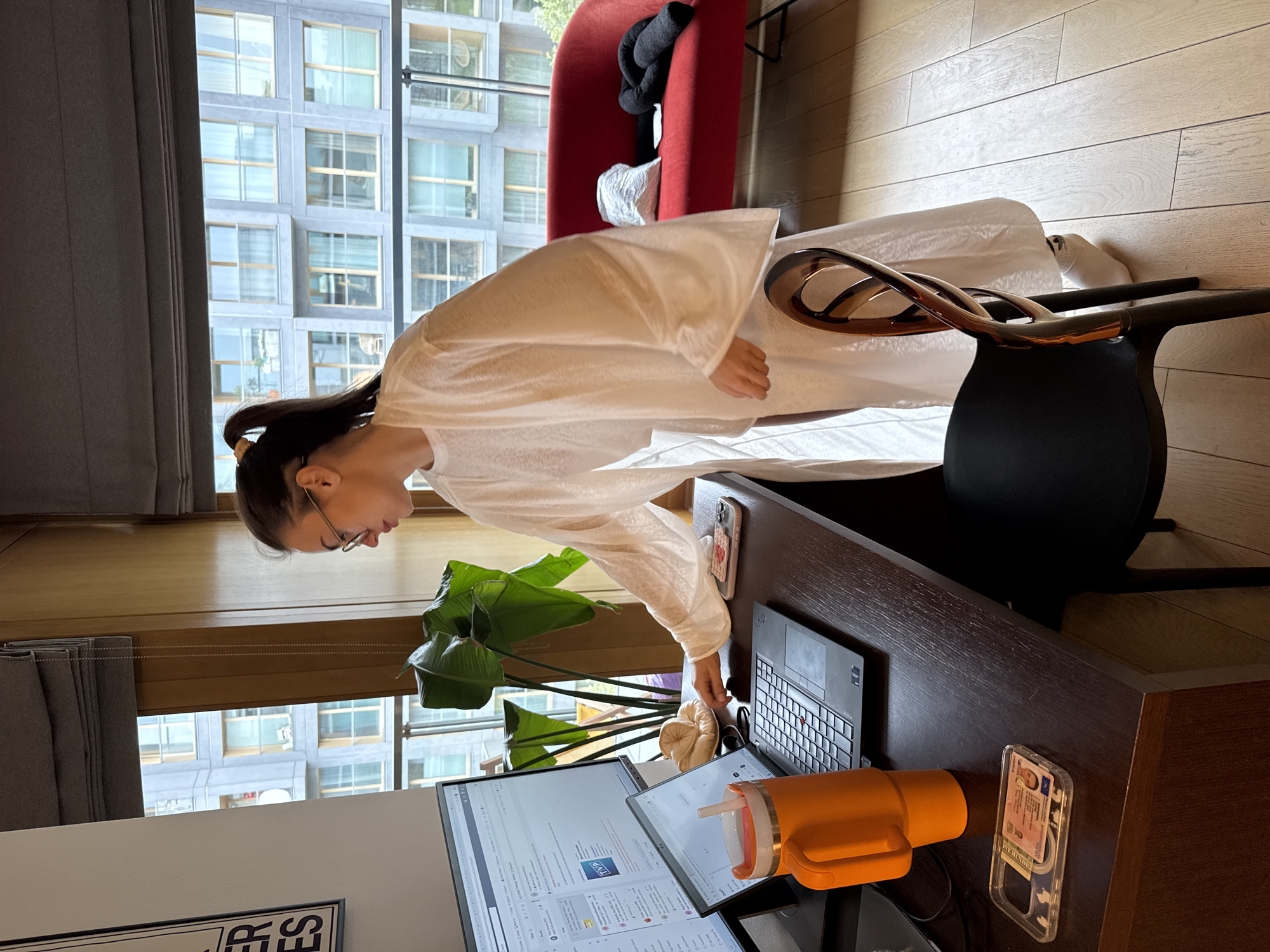 The image shows a woman in a white robe leaning over a desk in an interior setting. She has dark hair pulled back in a ponytail and wears glasses. Her hand hovers near a laptop on the desk.
The desk is made of dark wood and has two monitors, a tan-colored object, an orange water bottle, and a phone case with an identification card inside.
Behind the desk, there is a large window showing a cityscape of tall, modern buildings. A red chair is also visible near the window, and a black object sits on the chair. The floor is light wood. The city is Unknown, Unknown.