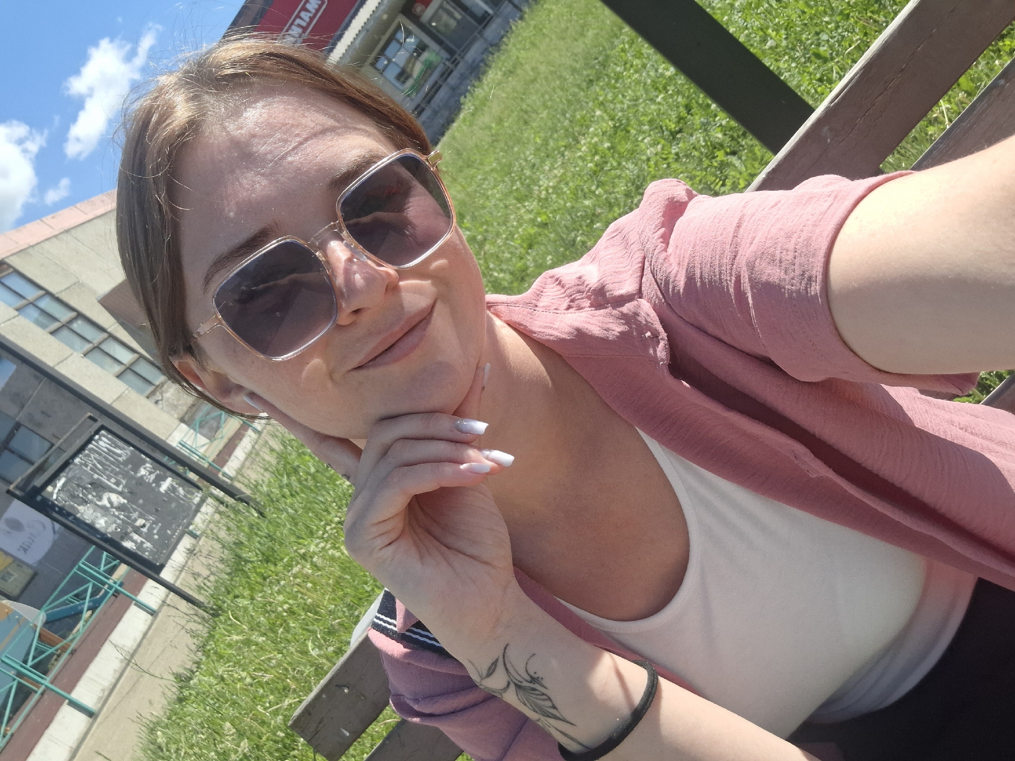 In the city of Markovsky, Russia, a woman is captured in a selfie, seated on a wooden bench outdoors. She wears oversized sunglasses and a pink open-front shirt over a white tank top. Her left arm rests on her chin, displaying manicured fingernails and a feather tattoo on her forearm, with a black band around her wrist. The background consists of lush green grass and a building under a clear blue sky with scattered clouds.