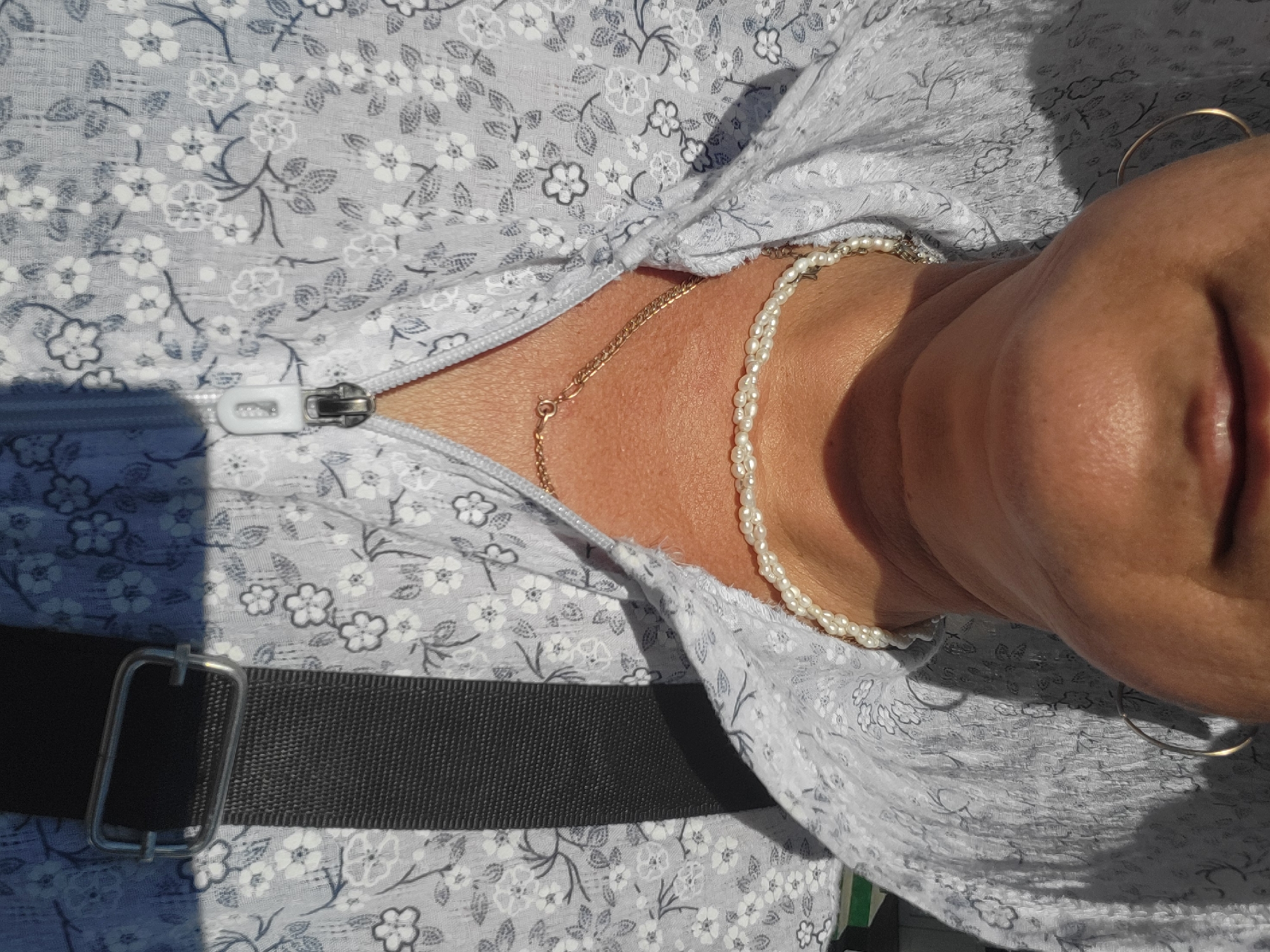 A medium close-up shot focuses on the neck and shoulders of a person wearing a light blue hooded jacket with a floral pattern. The jacket is zipped down, revealing a pearl necklace and a delicate gold chain. The person is also wearing a large gold hoop earring on their left ear. The background is blurred, but suggests an outdoor environment with sunlight casting shadows. There is no discernible connection to the city of Taldykorgan, Kazakhstan.