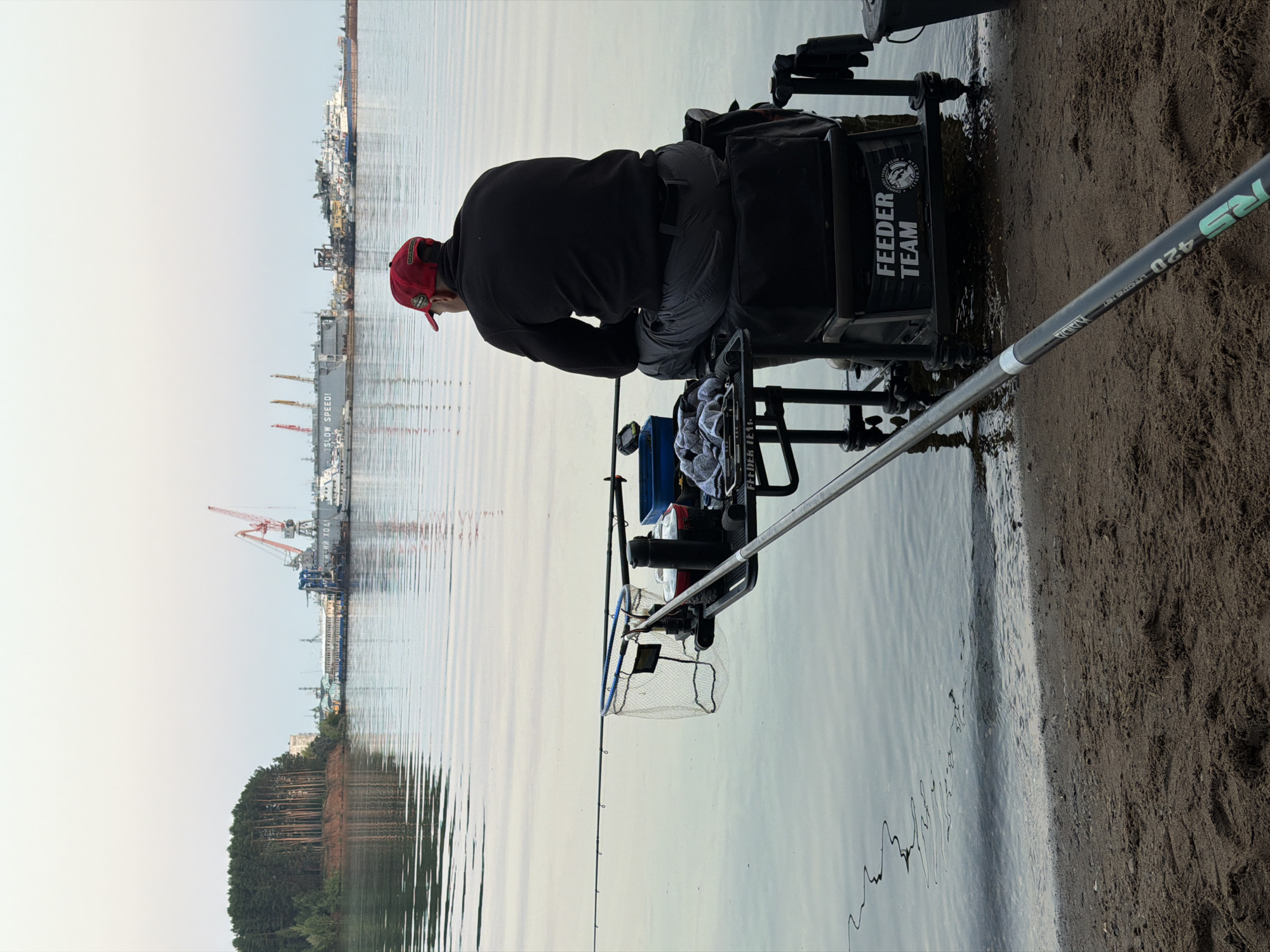 A lone fisherman, wearing a red cap and a black jacket, sits on a fishing seat next to a body of water. The fishing seat is equipped with various fishing gear, including rods, a net, and containers. In the background, a cityscape with industrial buildings and cranes is visible across the water. The scene is set on a sandy shore, with the water reflecting the sky and the distant structures. The image captures a moment of peaceful solitude by the water, with the industrial backdrop suggesting the proximity of an urban or industrial area. The specific location, Chaikovsky, Russia, can be inferred from the distinct industrial architecture and the overall environment, typical of Russian port cities.