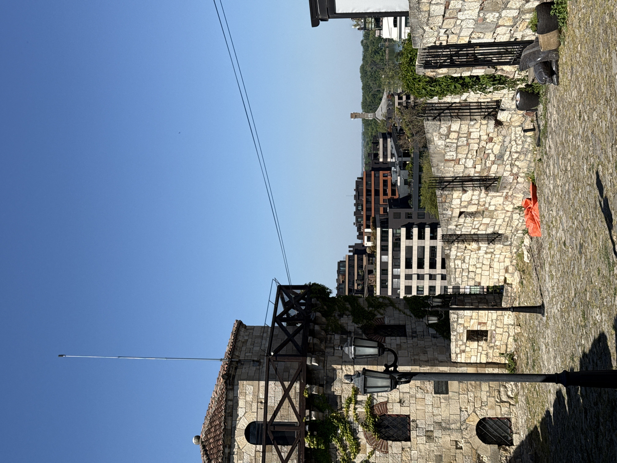 The image shows a view within the Belgrade Fortress in Belgrade, Serbia. It focuses on a stone wall structure, likely part of the old fortress walls, with metal support beams and ivy growing on the side. Several vintage-style street lamps stand in front of the wall. In the background, modern residential buildings can be seen, demonstrating the contrast between the historical fortress and the modern city. The foreground shows a cobblestone path. The sky is clear and blue. There are no people visible in the immediate scene. An orange tarp or fabric is visible on the wall.