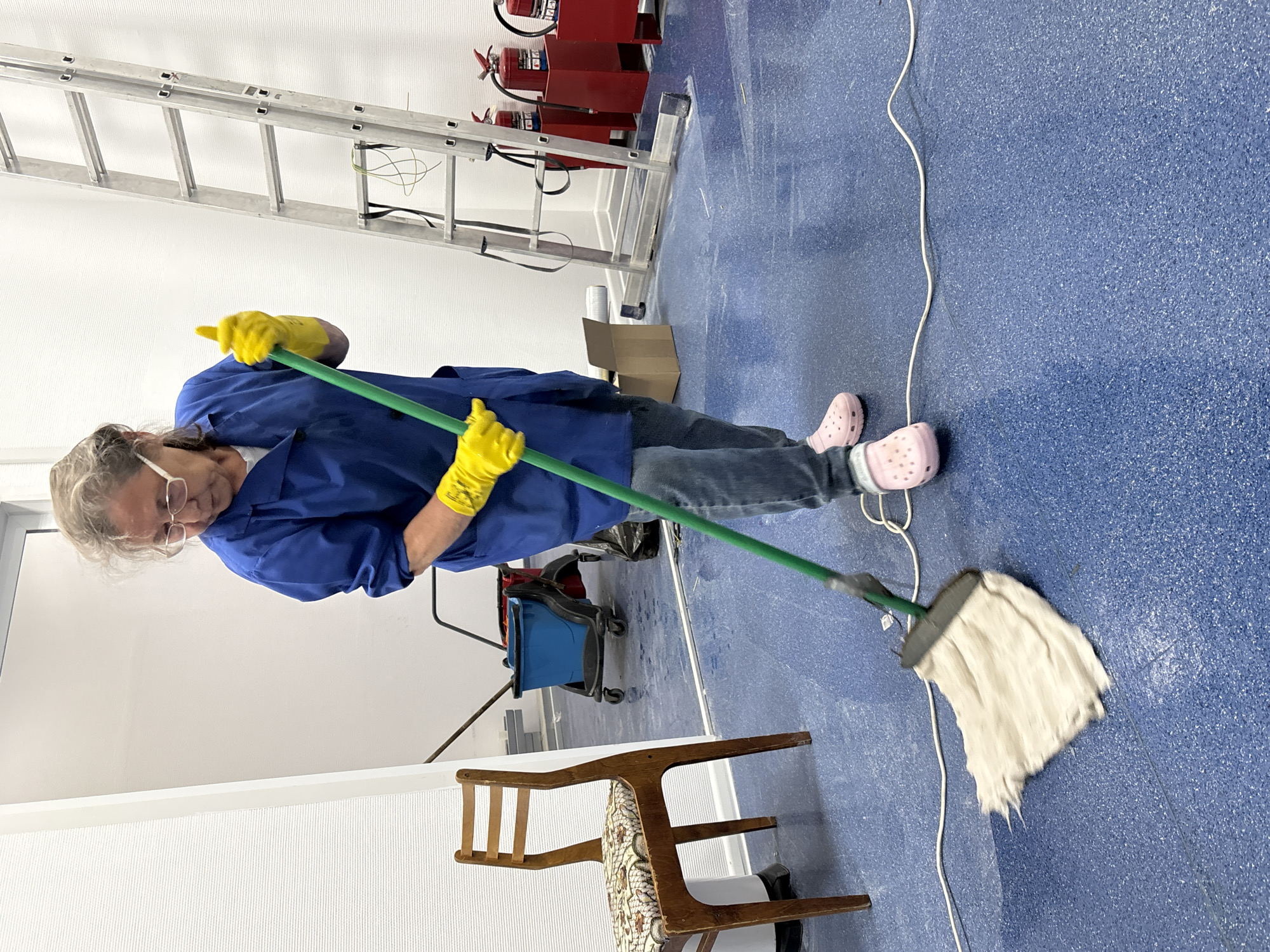 An elderly woman with short grey hair, wearing a blue button-down shirt, jeans, yellow rubber gloves, and pink Crocs, is mopping a blue speckled floor. She is holding a green-handled mop. To the left of the woman is a wooden chair with a patterned cushion, and further back is a blue bucket on wheels. In the background, a ladder and a fire extinguisher are visible against a white wall. A white electrical cord stretches across the floor. A small cardboard box is on the floor near the ladder. The location is not possible to determine with certainty as being in Izhevsk, Russia.
