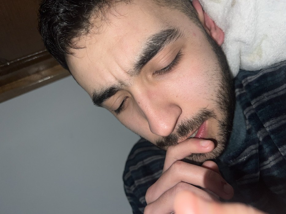 A close-up angled shot shows a man with dark hair, thick eyebrows, and a beard. His eyes are closed, and his brow is furrowed. He is lying on his side, with a white pillow or soft material near his ear. His fingers are near his mouth, and there is a blurred hand extending into the frame's bottom right. The background is a plain wall and part of a wooden structure. He appears thoughtful or concerned.