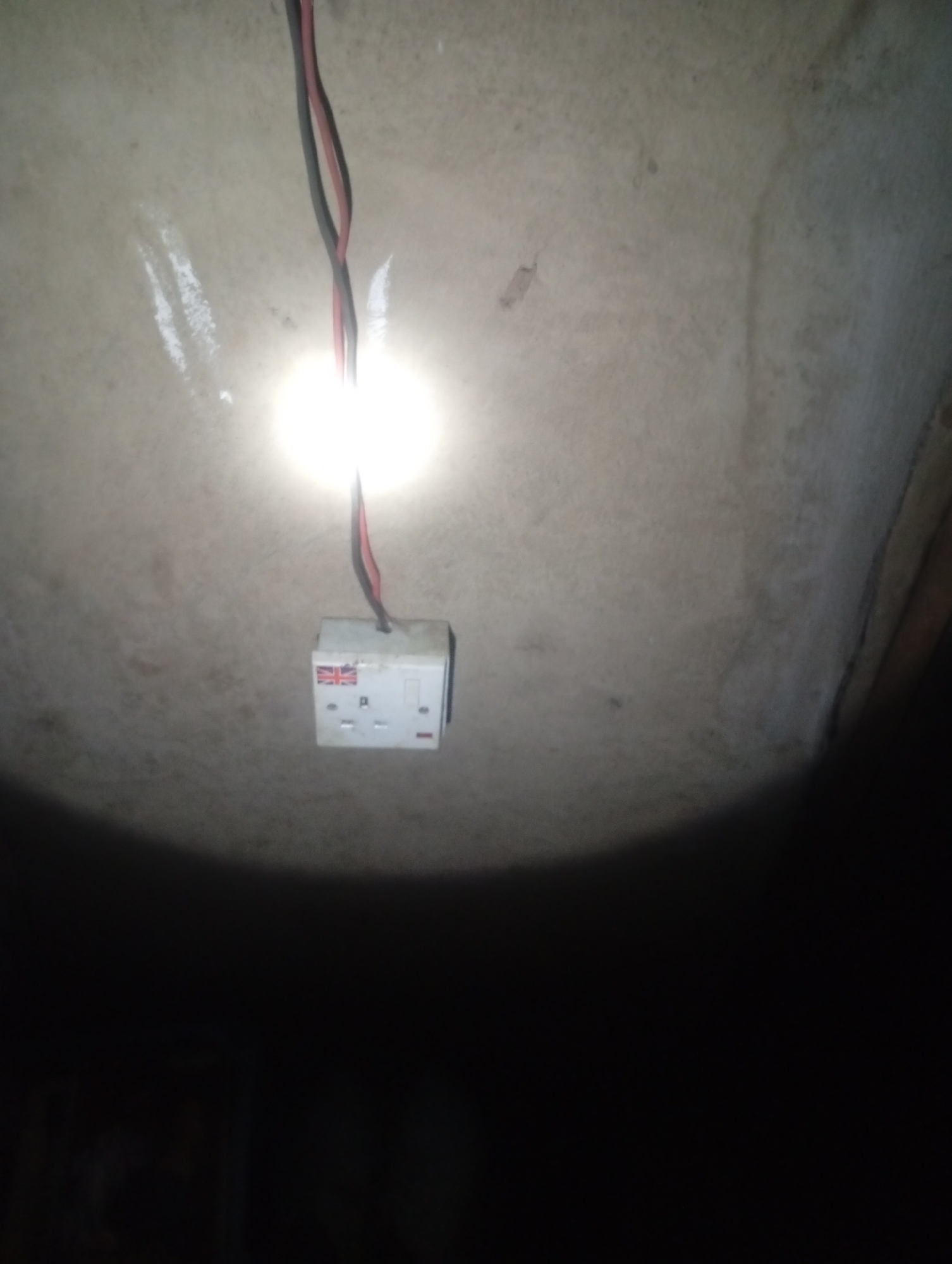 A white electrical socket with a Union Jack sticker is mounted on a textured, light-colored wall. Two electrical wires, one black and one red, hang from the top of the socket. A bright light source illuminates the socket and a portion of the surrounding wall. The rest of the image is dark, with the bottom portion obscured by shadow. The scene is likely an indoor electrical installation. The location in Jalingo, Nigeria, cannot be determined from the visual information alone.