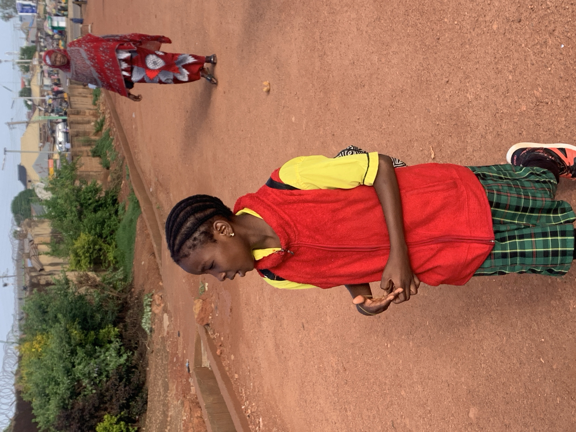 In Jos, Nigeria, on a dirt road, a young Black girl with braided hair is kneeling on the ground. She is wearing a yellow shirt, a red vest, and a green plaid skirt. Her hands are clasped in front of her. In the background, a Black woman in a red and white patterned headscarf and dress is walking away from the camera. Other people and buildings are visible in the distance, along with lush green vegetation and a barbed-wire fence. The overall impression is a busy, dusty street scene in a Nigerian city.