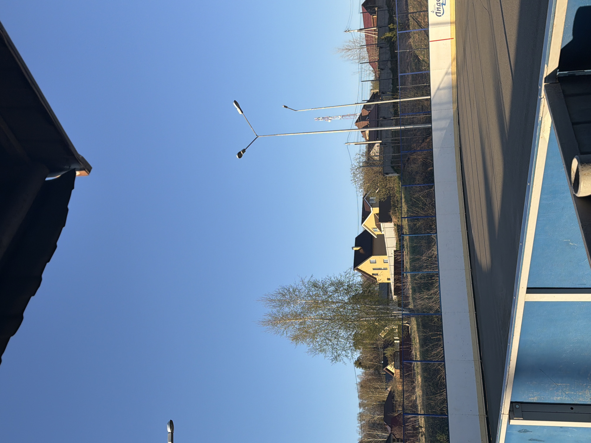 The image depicts an outdoor scene in Noviy, Russia, under a clear blue sky. A portion of a dark, possibly metal, roof is visible in the upper-left corner. Adjacent to it are street lights. Below the streetlights are houses, a yellow house is visible in the center, surrounded by bare trees and other vegetation. To the right is a sports court, defined by a white wall, and blue fencing. The visible court surface is dark grey. Part of an overhead shelter made of blue materials is in the upper-right corner.