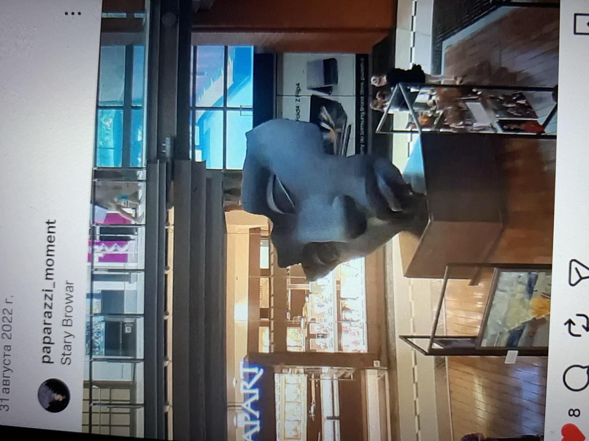 The image depicts a large, abstract sculpture of a face, rendered in a gray, matte material. The sculpture is positioned in a well-lit interior space, likely a shopping mall or public atrium, indicated by the presence of storefronts in the background, one of which has a neon sign reading "APART." The sculpture sits on a dark wooden pedestal. In the background, several individuals are visible, including a group of people standing near a railing, and a person in a black dress on the right side of the frame. The overall environment suggests a commercial or entertainment venue. The provided text at the top of the image, "31 августа 2022 г.", indicates the date. The username "paparazzi_moment" and the location "Stary Browar" are also displayed.