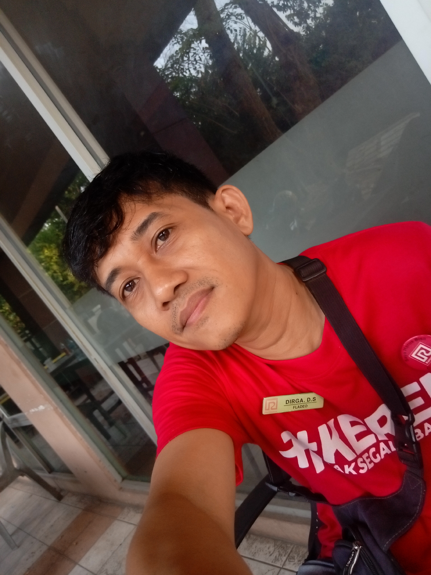 A man with dark hair and a mustache is taking a selfie in front of a window that reflects trees and sky. He is wearing a red t-shirt with white text that appears to say "KER...BAK SEGA..." and a badge that reads "DIRGA. D.S FLADEO". He also has a black strap across his chest and a dark bag. The flooring appears to be tiled. The lighting suggests it is daytime. The style of the building and the greenery visible in the reflection are consistent with locations in Batam, Indonesia, possibly in an outdoor seating area of a restaurant or cafe.