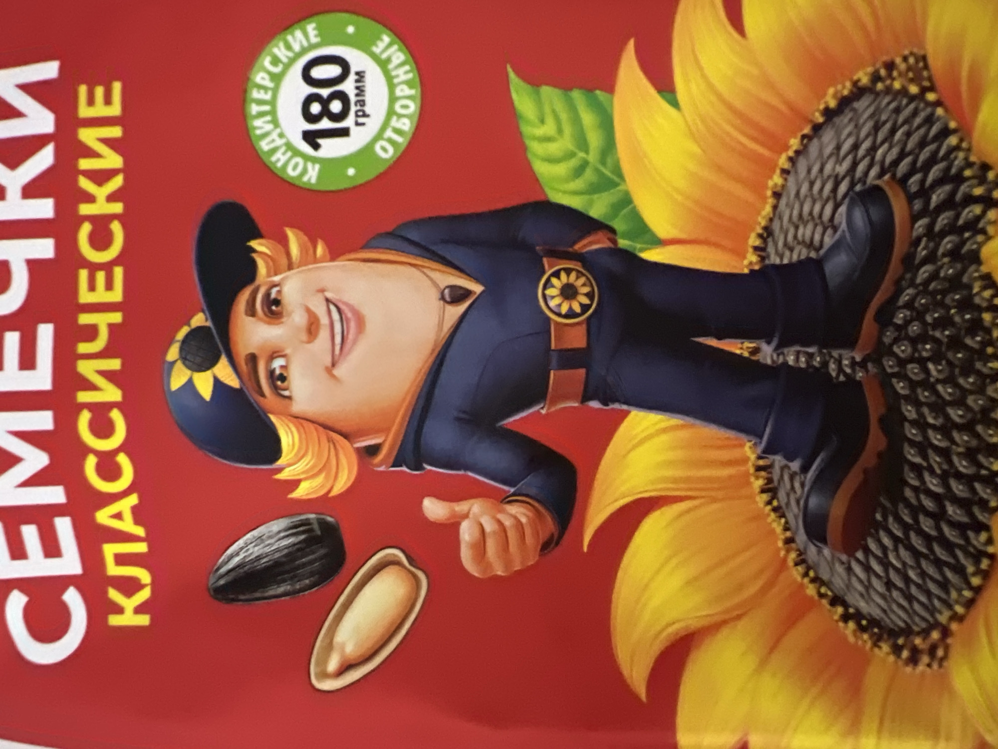 The image displays a product packaging, likely for sunflower seeds, set against a solid red background. Prominently featured at the top are two lines of text: "СЕМЕЧКИ" (SEMECHKI), in large white capital letters, followed by "КЛАССИЧЕСКИЕ" (KLASSICHESKIYE), in smaller yellow capital letters, which translates to "Sunflower Seeds, Classic."
The central element is a smiling anthropomorphic character with an oversized, round, light-skinned face and a smaller body, resembling a humanized sunflower seed. The character wears a dark blue baseball cap adorned with a yellow sunflower symbol and has curly blond hair extending from beneath the cap. Dressed in a dark blue jumpsuit or tracksuit, the character features a brown belt with a large yellow sunflower buckle and a dark pendant around the neck. Dark brown boots with light brown soles are visible. The character's right hand (viewer's left) is raised in a thumbs-up gesture.
To the upper left of the character, a whole black sunflower seed floats above an open, light-colored sunflower kernel resting in a small, pale tray or shell fragment.
The character stands atop a large, vibrant yellow sunflower with a dark, textured center. A single green leaf is partially visible behind the character's right shoulder (viewer's left).
On the right side of the image, there is a circular green emblem with a white border. Inside the emblem, white Cyrillic text reads "КОНДИТЕРСКИЕ" (KONDITERSKIYE - Confectionery) curved along the top and "ОТБОРНЫЕ" (OTBORNYE - Selected) curved along the bottom. The center displays large white numbers "180" above the word "грамм" (gramm - grams).
The image provides no visual or textual information regarding its location in the city of Sochi, Russia.