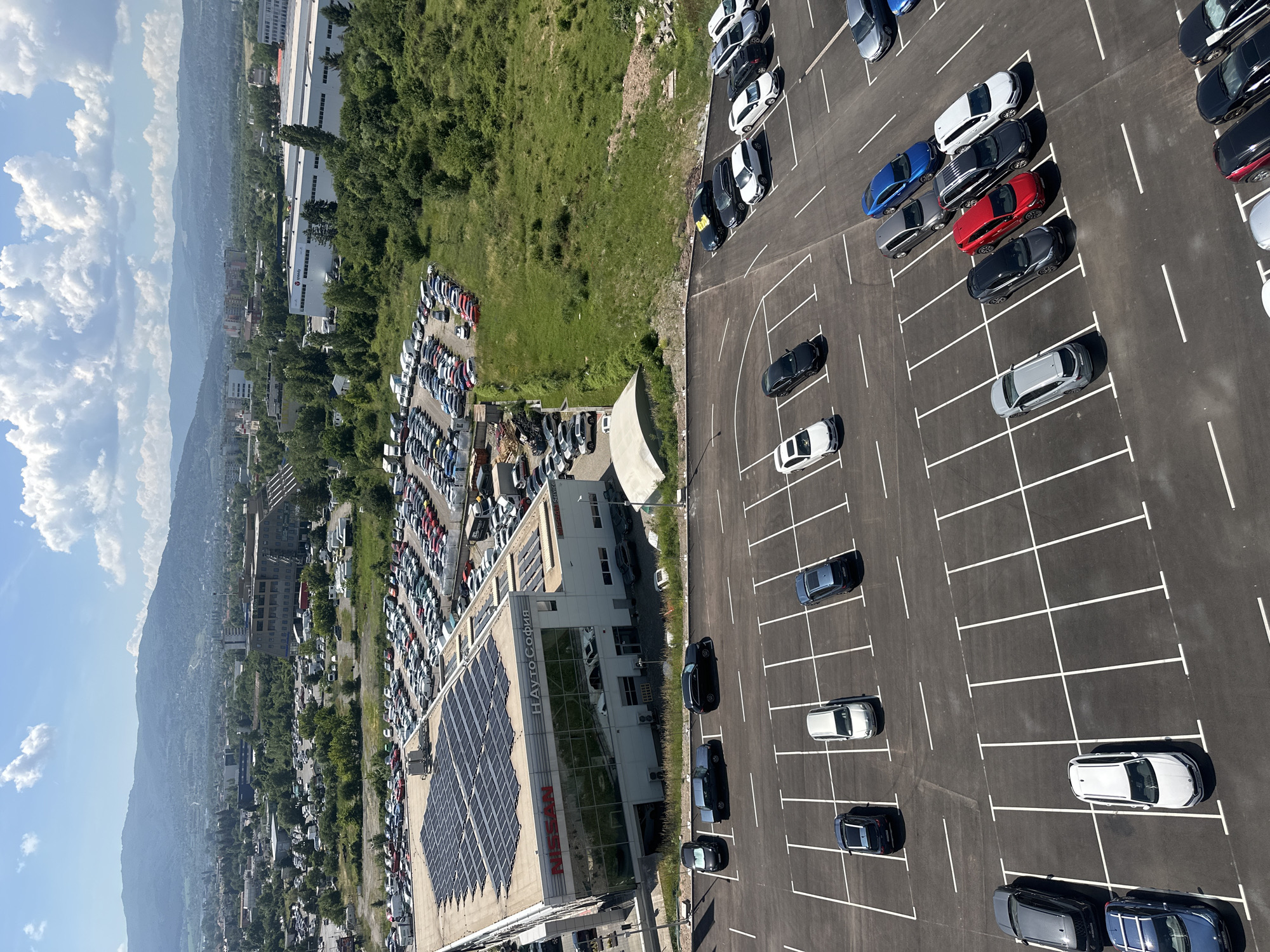 This is a high-angle aerial photograph of a car dealership and its surrounding area in Sofia, Bulgaria. The primary subject is a modern building with a large solar panel array on its roof, identified by "NISSAN" and "НАУТО СОФИЯ" signage. The building has a large glass facade reflecting the sky and interior car displays. Adjacent to the building is a vast parking lot filled with numerous cars, both new and used, organized in marked bays.
Beyond the dealership, the scene transitions to a more urban and then a natural landscape. In the mid-ground, there are more buildings and industrial-looking structures, interspersed with trees and greenery. Further in the distance, a city skyline with buildings of varying heights is visible, set against a backdrop of rolling green hills and mountains under a clear blue sky with scattered white clouds.
The foreground of the image focuses on a meticulously organized parking lot, with rows of cars parked in designated spots. The ground is asphalt, marked with white lines indicating parking spaces. Several cars are clearly visible in this lot, displaying a variety of makes and models in different colors. The lighting suggests it is daytime, with sunlight casting shadows from the cars and building. There are no people visible in the photograph. The environment appears to be well-maintained and organized, typical of a commercial car sales and service center.
