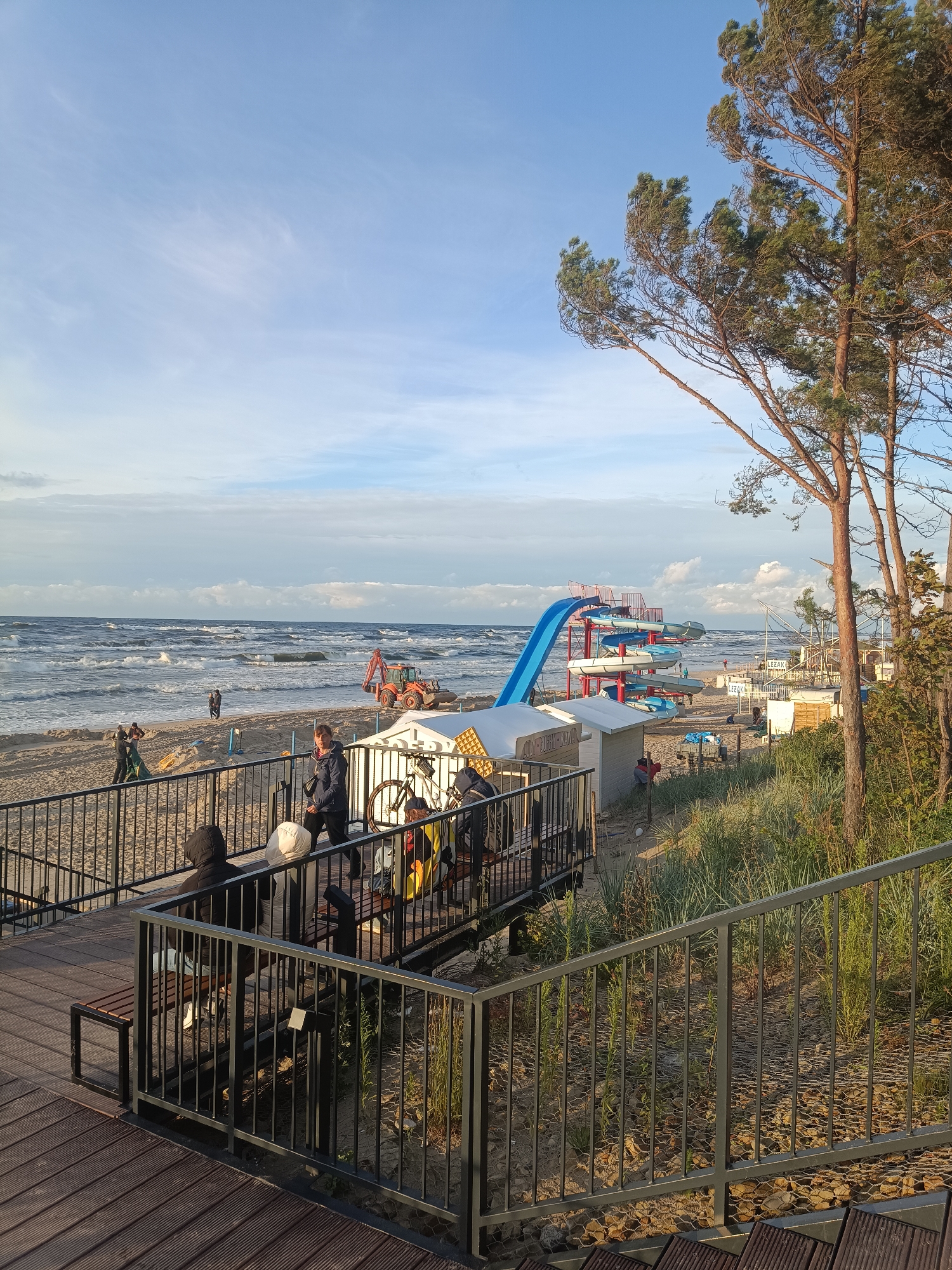 This media file captures a beach scene in Stegna, Poland. The time of day appears to be late afternoon, as indicated by the warm, low-angled sunlight illuminating the scene and the shadows cast by the trees. The weather is partly cloudy with a gentle breeze creating waves on the sea.
The scene depicts a beach resort area with a prominent blue water slide structure, a red excavator on the sandy shore, and several small white buildings that might be concessions or changing rooms. In the foreground, a wooden boardwalk with metal railings leads to a viewing area. Several people are visible on the boardwalk, some sitting on benches, while others are standing and observing the beach and sea.
On the beach, a few individuals are scattered, with one person in the middle ground appearing to be interacting with a green tarp. Further down the beach, the excavator is parked near the water's edge. The sea is a bit choppy, with whitecaps visible on the waves.
To the right of the frame, tall pine trees with sparse green needles rise against the sky, adding a natural element to the developed resort setting. The overall atmosphere is one of a relaxed, end-of-day leisure activity at a seaside location.
Visible text includes "F.ZAK." on a white building in the middle ground.