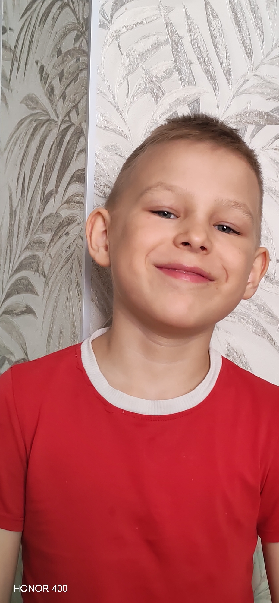 A young boy with short, light-colored hair smiles at the camera. He is wearing a red t-shirt with a white collar. The background consists of a patterned wallpaper with leaf-like designs. The location is in Noviy, Russia, based on the 'HONOR 400' watermark in the lower left of the frame.