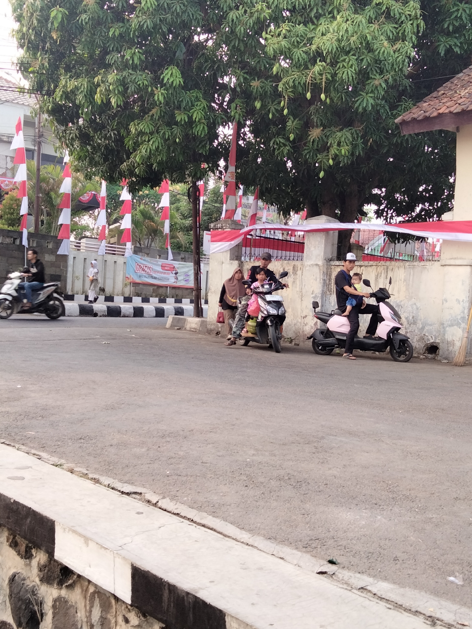 The image depicts a street scene in Cariu, Indonesia, adorned with red and white Indonesian flags. On the left, a person rides a white motorcycle. Further down the street, several individuals are gathered around two scooters. One scooter, a dark color, carries at least two people. Another scooter, pastel pink, is ridden by a man holding a baby on his chest. The background features a wall with a banner, a large tree with visible fruit, and buildings. The street itself appears to be asphalt, with a concrete curb marked with black and white stripes.