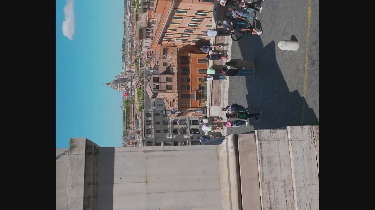 The image shows a wide shot of a European city, likely Rome, given the architectural style and the dome of a prominent church in the background. The foreground features a large stone structure on the left, casting a shadow. In the middle ground, a paved plaza is populated with numerous people walking, standing, and conversing. Some individuals are dressed casually, suggesting a tourist area. There are buildings with terracotta roofs and various architectural details, typical of older European cities. A white cylindrical bollard is visible on the plaza. The sky is bright blue with a few scattered clouds. The overall scene depicts a sunny day in an urban environment with a significant amount of pedestrian activity. The reference to Rignano Flaminio, Italy is not supported by any visual evidence within the image itself; the architecture and cityscape are more consistent with a major metropolitan center like Rome.