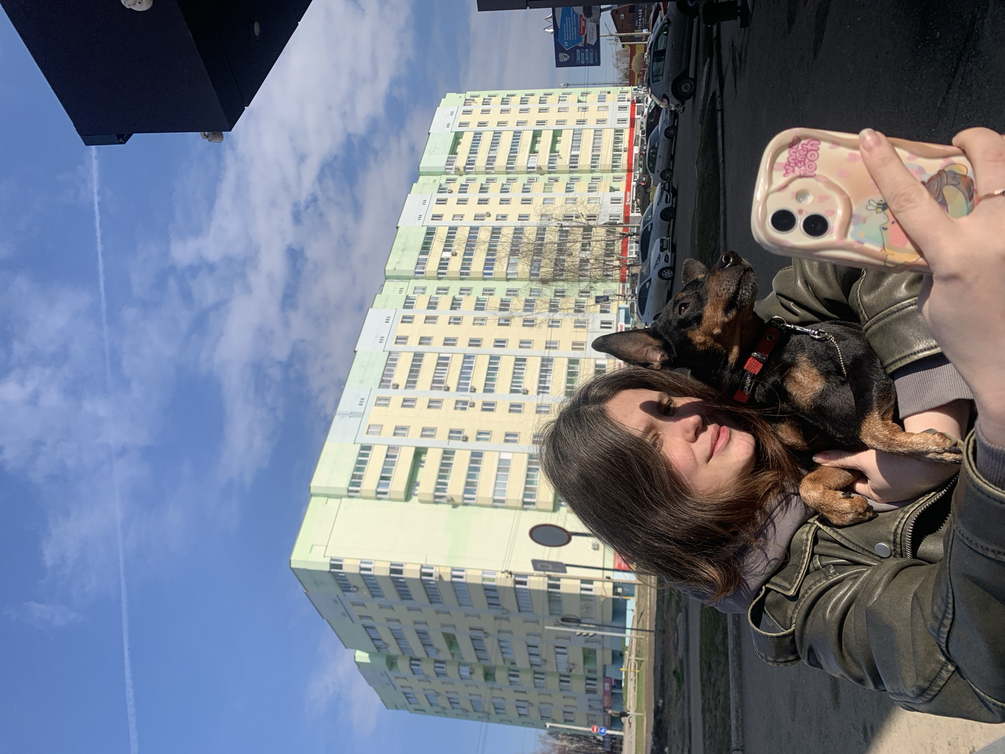 A young woman is taking a selfie outdoors with her small dog. The image is taken from a slightly low angle, showing the upper part of her body and the dog she is holding. She has brown hair and is wearing a dark olive green leather jacket over a grey hooded sweatshirt. Her left hand holds a smartphone with a light-colored, patterned case featuring Winnie the Pooh characters and partially visible text "WORRY HAPPY".
Cradled in her arms is a small, dark-colored dog, possibly a Miniature Pinscher, with tan markings on its face and paws. The dog wears a red collar with some unreadable text and is looking upwards and slightly to the left. The woman has a calm, pleasant expression with a subtle smile.
In the background, under a bright blue sky with scattered white clouds and a visible airplane contrail, stand large, multi-story residential buildings. These buildings feature light yellow facades with light green sections and numerous windows, many equipped with air conditioning units. A street with parked cars is visible to the right of the buildings. The scene depicts a sunny day in an urban environment, characteristic of a city like Chaikovsky, Russia.