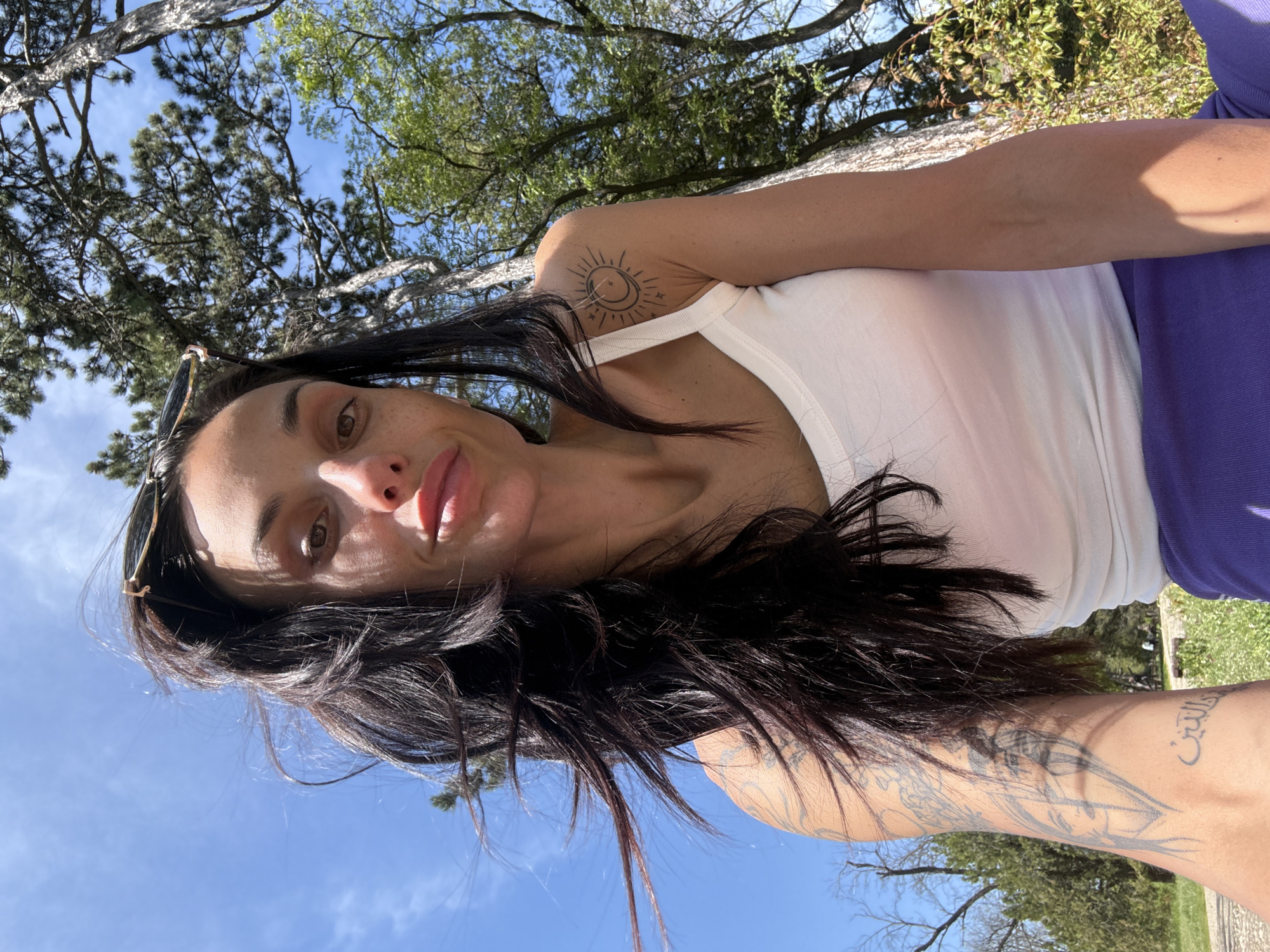 This rotated, low-angle, selfie-style photograph captures a woman outdoors under a bright blue sky. She has long, dark hair and is looking directly at the camera with a relaxed expression, her lips slightly parted. Sunglasses are pushed up onto her head. She is wearing a white tank top and a purple garment on her lower body.
Two distinct tattoos are visible: a stylized sun and crescent moon design with small stars is on her left upper arm/shoulder. On her right forearm, there is a larger, intricate line-art tattoo, possibly featuring figures or abstract elements, alongside some visible Arabic script.
The scene is set outdoors amidst various trees, both coniferous and deciduous, suggesting a park or natural green space. The bright sunlight and clear blue sky indicate it is daytime. The image captures a moment of leisure or self-portraiture in an outdoor setting, likely in Pleven, Bulgaria, though no specific geographical landmarks are discernible from the image itself.
