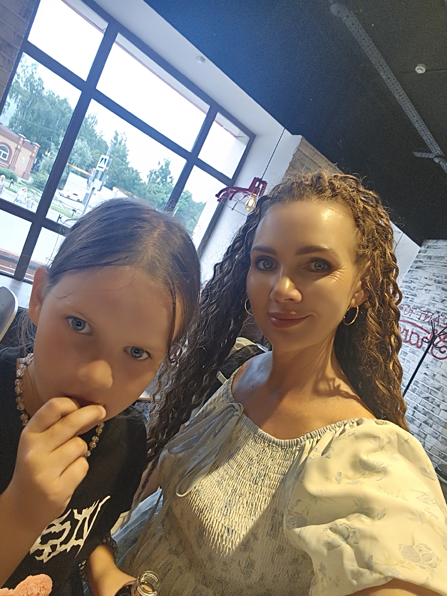 The image is a selfie taken indoors, likely in a cafe or restaurant, with a large window in the background that overlooks a street scene in the city of Chaikovsky, Russia. Through the window, a brick building with a rounded archway, trees, and a traffic sign are visible. The interior appears to have exposed brick walls and a dark ceiling with industrial-style lighting.
In the foreground, two individuals are captured. On the left, a young girl with dark hair is looking towards the camera, her hand near her mouth. She is wearing a black t-shirt with a white graphic design and a beaded necklace. To the right of the girl is a woman with long, curly brown hair, wearing a light-colored dress with a floral pattern. She is looking at the camera and smiling. Her attire includes hoop earrings. The overall lighting suggests an indoor setting with natural light coming from the window.