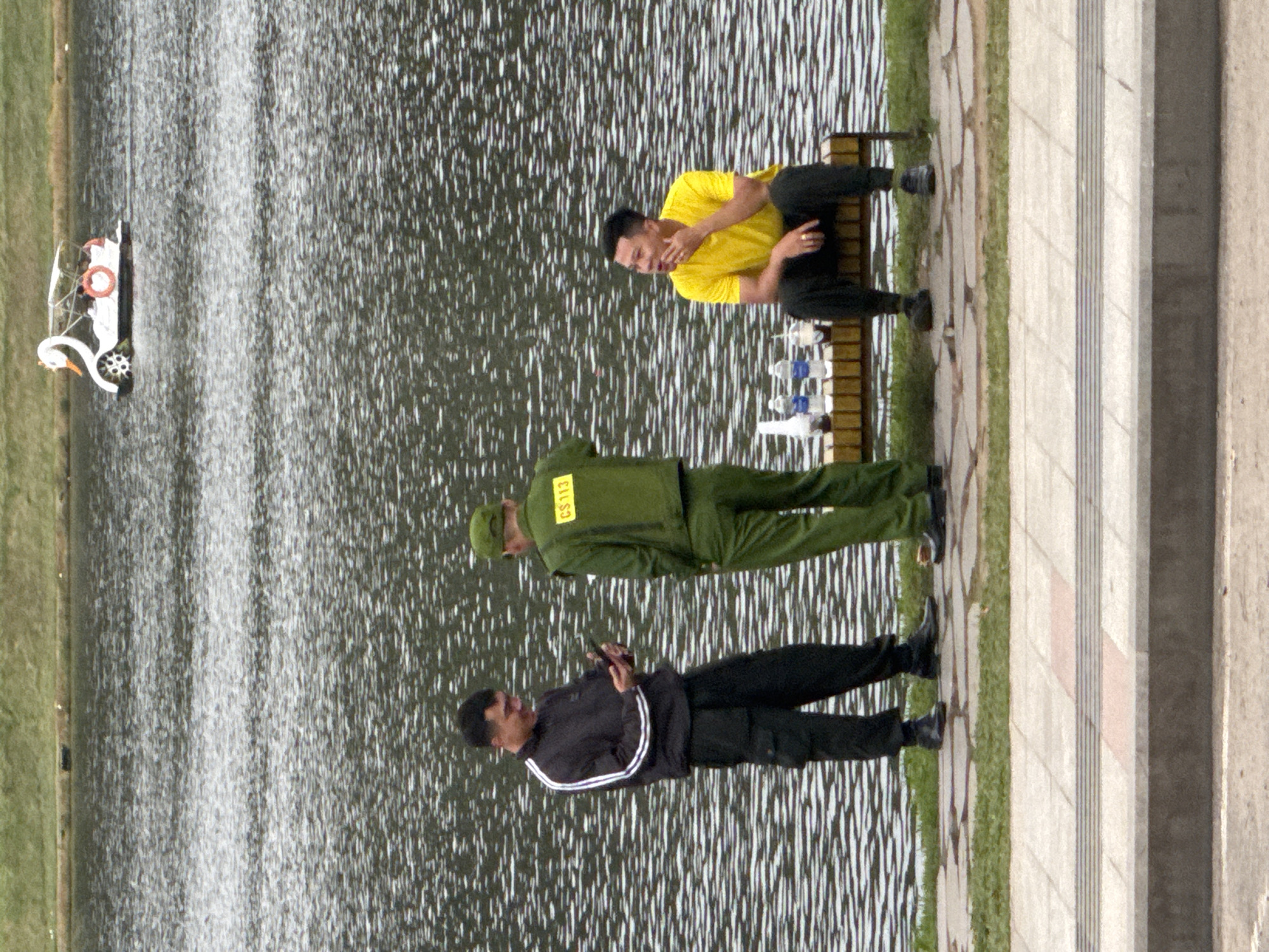 In Da Lat, Vietnam, three men are positioned near a body of water. On the left, a man in a black jacket with white stripes and black pants is holding a phone. In the center, a man in a green uniform with the insignia "CS 113" stands facing away from the camera, looking at the man on his left. To the right, a man in a yellow t-shirt and black pants is seated on a wooden bench, looking to his left with his hand raised to his chin. The bench also holds three bottles of water. In the background, a swan-shaped paddle boat is visible on the water. The environment includes a grassy bank and a paved walkway.