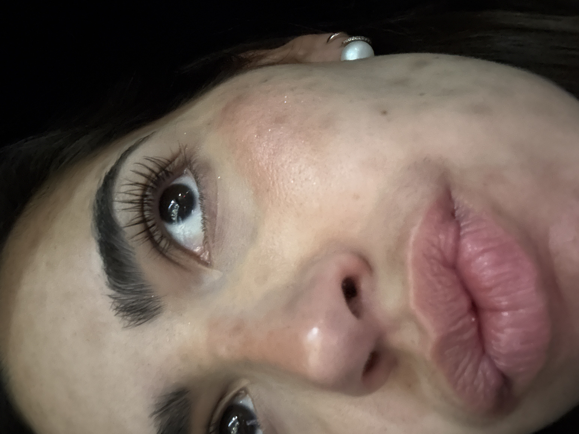 A close-up, upward-angled view of a woman's face. She has dark hair, light skin, and prominent eyelashes. Her lips are full and tinted pink. A ring, featuring a white pearl, is visible above her forehead. The background is dark and indistinct. There's no information to locate this image in San Bernardino Tlaxcalancingo, Mexico.