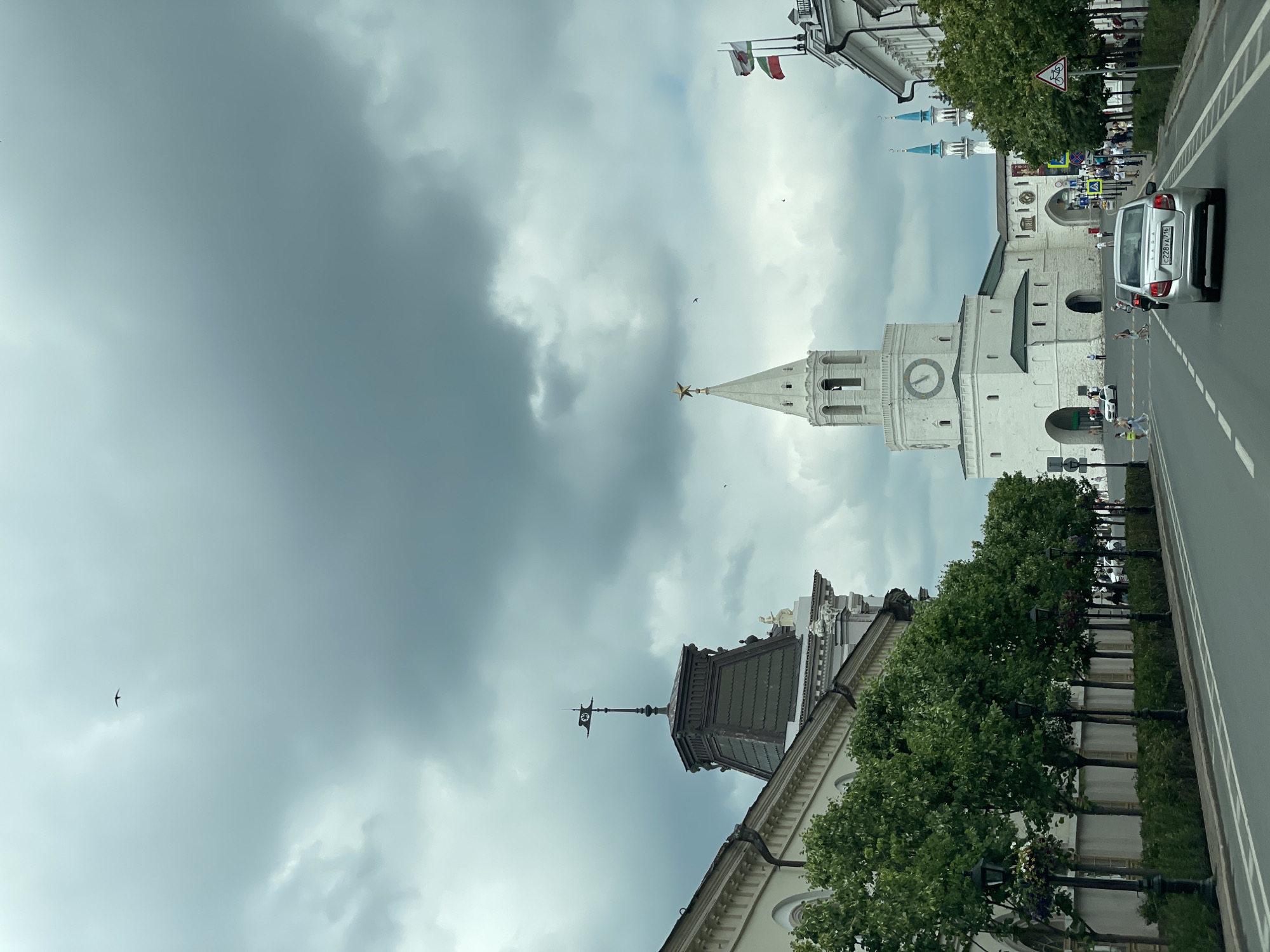 The image shows a view of the Kazan Kremlin in Kazan, Russia, under a cloudy sky. In the foreground, a silver Lada car is driving down a street lined with trees. On the left, a white building with an ornate roof and a weather vane is visible. To the right of the street, a prominent white bell tower with a clock face and a golden star at its peak stands tall. Further in the background, blue minarets of a mosque can be seen. Several people are walking on the sidewalks and crossing the street. The overall scene depicts a historical urban environment with a sense of movement and daily life.