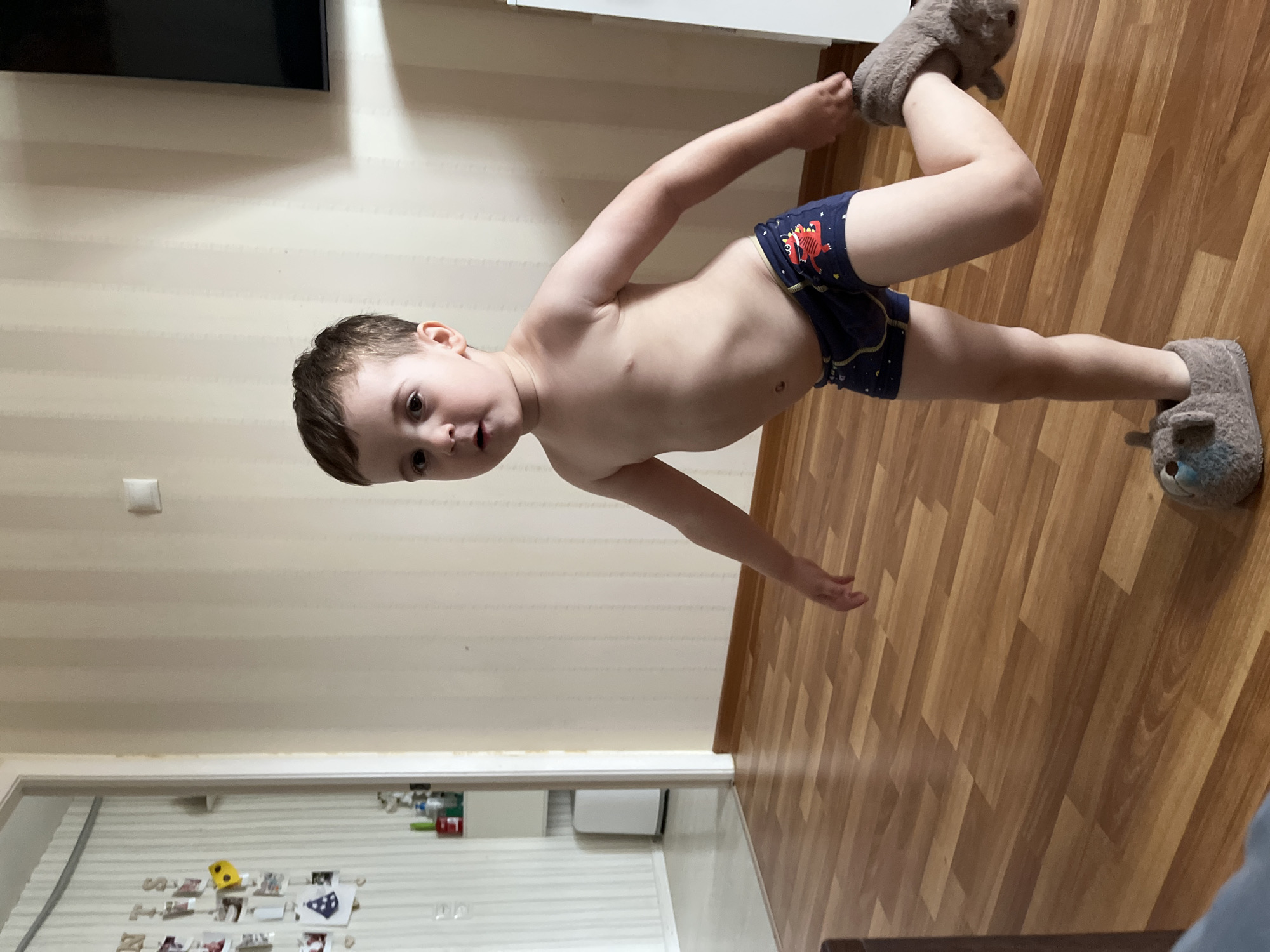A young male toddler, estimated 2-4 years old, stands indoors in a residence located in Chaikovsky, Russia. The child has short, dark brown hair and dark eyes, looking directly at the camera with a neutral expression and slightly parted lips. He is shirtless and wears dark blue boxer briefs patterned with red cartoon dinosaurs. On his feet are grey, animal-shaped slippers; the child's left foot is firmly planted, while his right foot is lifted, bent at the knee, and partially dislodged from its slipper.
The flooring consists of light brown laminate wood planks. The walls are covered in light beige wallpaper with subtle vertical stripes. To the left, a white doorframe leads to an adjacent room, also with striped wallpaper. Within this background room, a white cabinet or shelf holds various bottles, and a tall, white, narrow appliance stands on the floor. On the wall beside the doorway, a string or wire displays multiple small, pinned photographs or notes, with visible letters partially forming "N T S" and including yellow and blue decorative elements. On the right-hand wall, a white light switch is visible, and the upper right portion of a wall-mounted, dark television screen can be seen. The edge of a white piece of furniture is also present on the far right of the image.