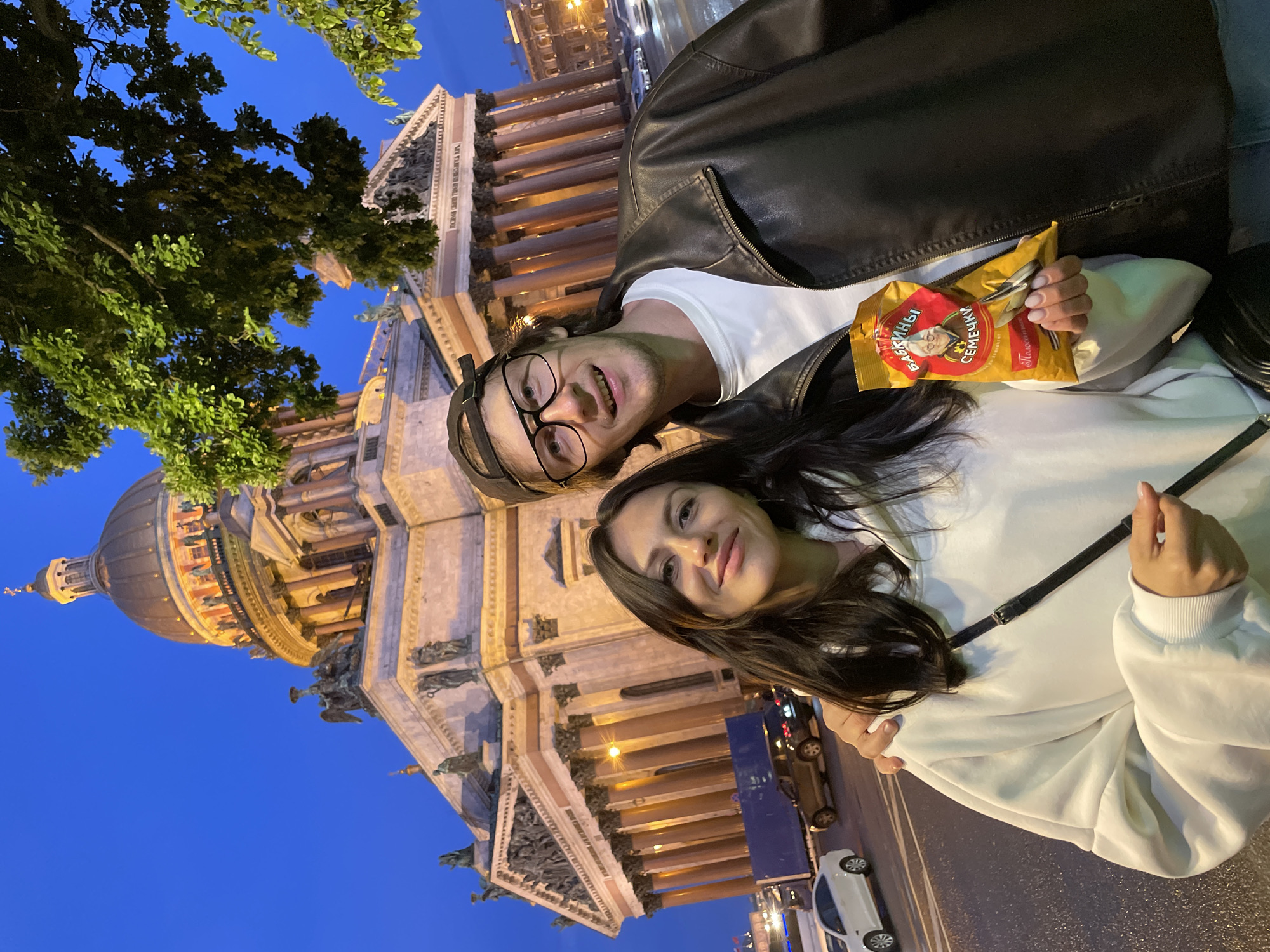 A young couple poses for a selfie in front of Saint Isaac's Cathedral in Saint Petersburg, Russia, at dusk. The woman, with long dark hair, wears a white hooded sweatshirt and a black crossbody bag. She smiles at the camera. The man, wearing glasses, a black baseball cap, and a black leather jacket over a white t-shirt, stands beside her. He also smiles. The woman holds a bag of sunflower seeds. The cathedral's dome and facade are illuminated, with its columns and statues clearly visible against the blue evening sky. Trees overhang the scene from the right. Cars and streetlights are visible in the background on a wet street.