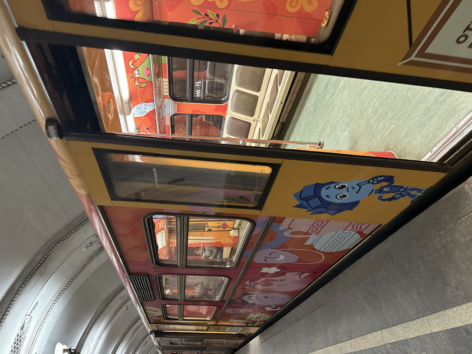 The image captures a brightly decorated subway train traveling through a station. The train's exterior is adorned with cartoonish illustrations and a color palette dominated by oranges, yellows, and blues. The tunnel's wall, adjacent to the train, is constructed from light gray concrete with a curved, ribbed design. Through the train's windows, glimpses of the interior can be seen, including passengers and seating. The reflection in the windows indicates another train traveling in the opposite direction.