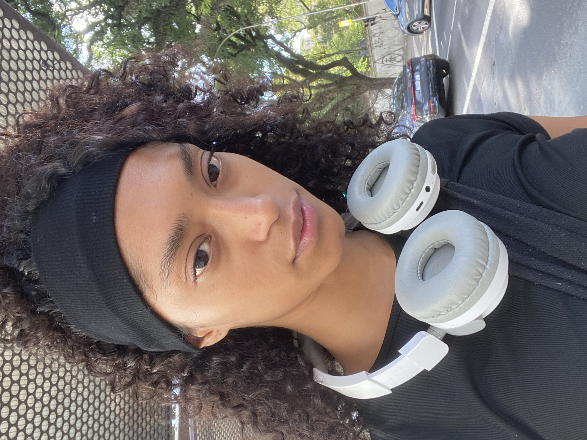 A close-up, slightly rotated, eye-level view of a young woman with brown curly hair wearing a black headband and gray over-ear headphones around her neck. She is wearing a black shirt and looking directly at the camera. A perforated metal surface is visible on the left side of the frame. Behind her, out-of-focus, are trees and a street scene. Based on visual clues such as vegetation and building style, this image was likely taken in the city of São Paulo, Brazil. A black car and other parked vehicles are visible in the background street scene.